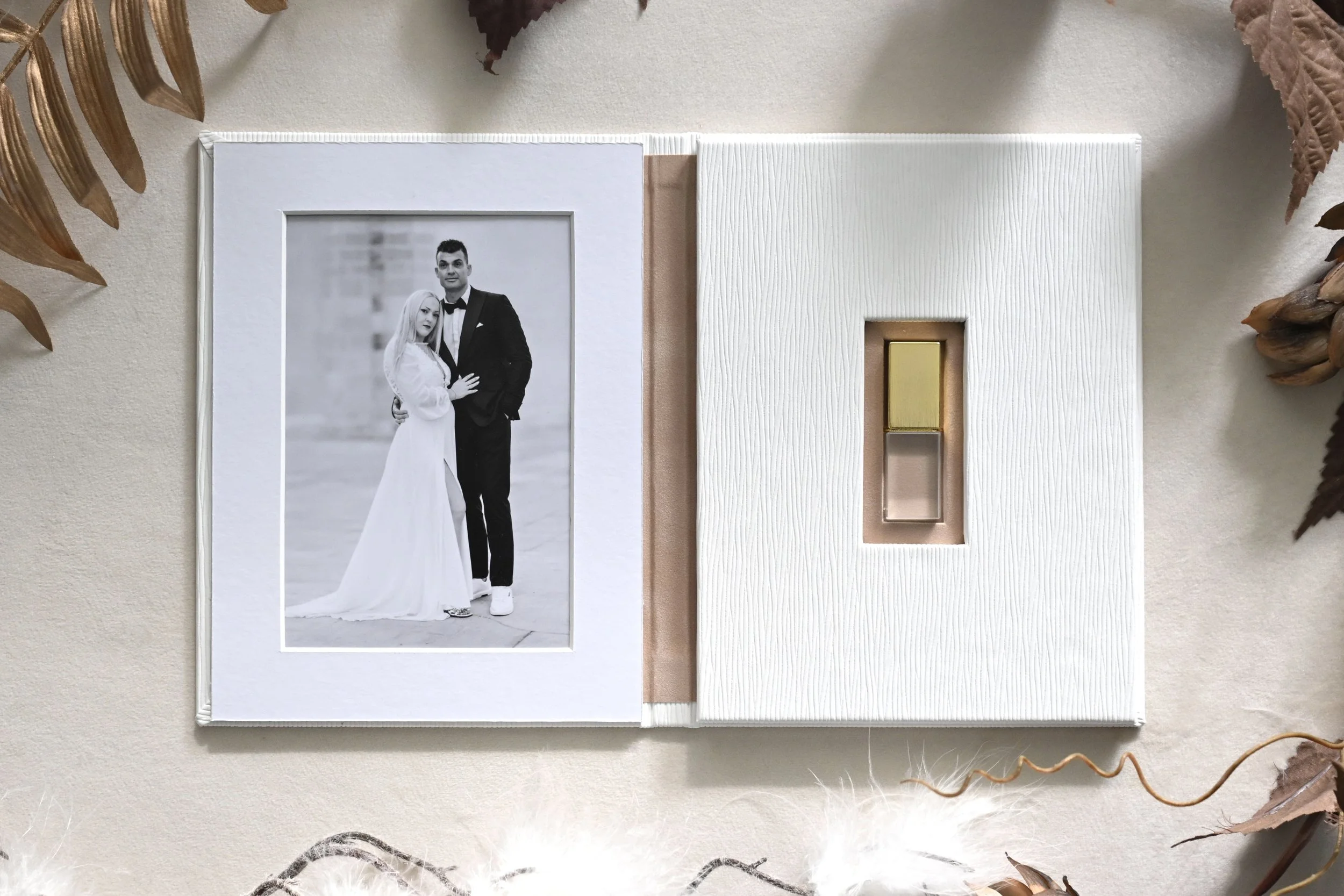 A black-and-white photograph of a couple in wedding attire inside a decorative luxury usb folio, with a light-colored textured fabric background and surrounded by dried leaves and natural elements.