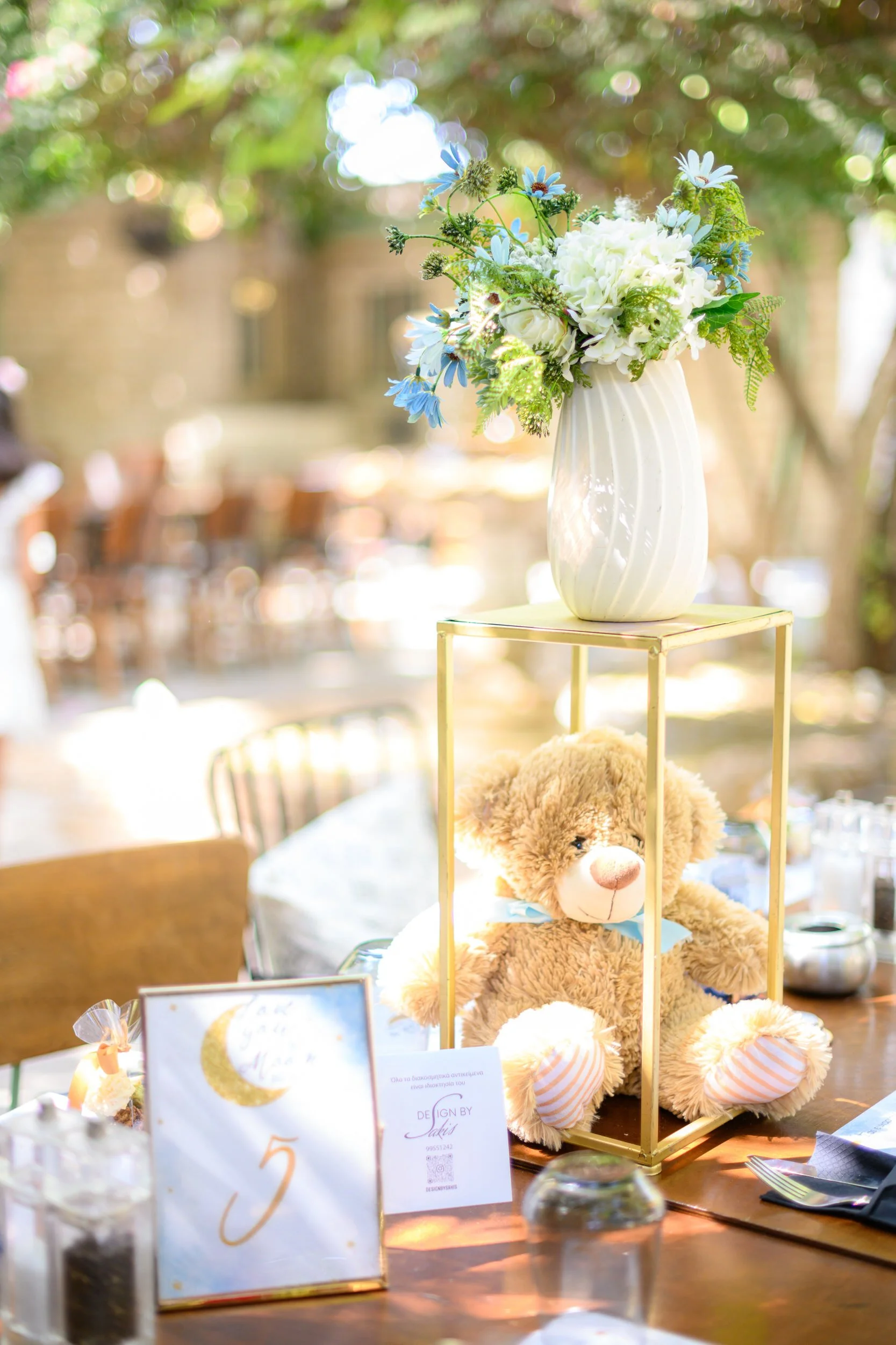 A table centerpiece with a white vase holding a bouquet of white and blue flowers, a stuffed teddy bear with a blue ribbon inside a gold cage, and a table number sign with a moon and star illustration for a wedding or event.