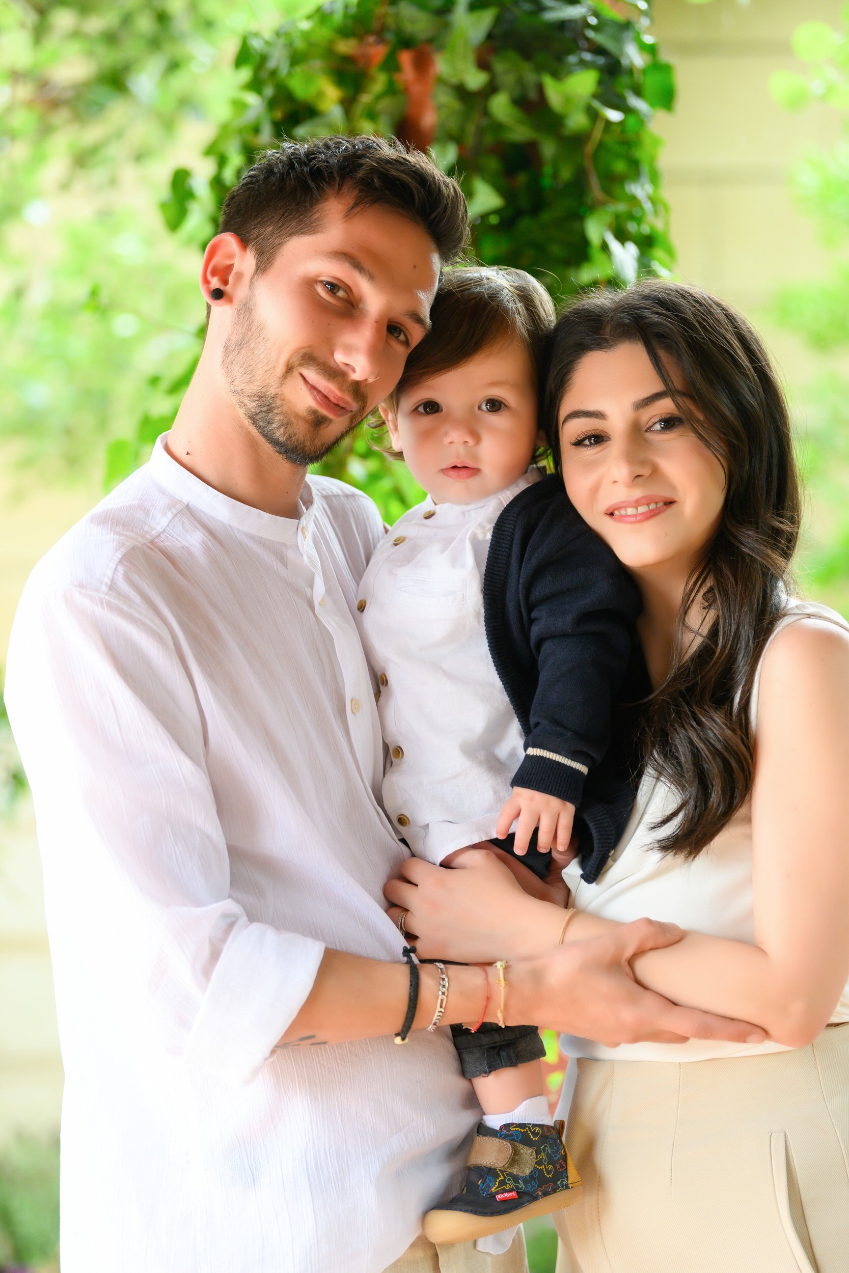 A happy family portrait outdoors featuring a man, woman, and a young child. The man has short dark hair, a beard, and wears a white shirt. The woman has long dark hair and wears a white top. The child has short dark hair, is dressed in a white shirt 