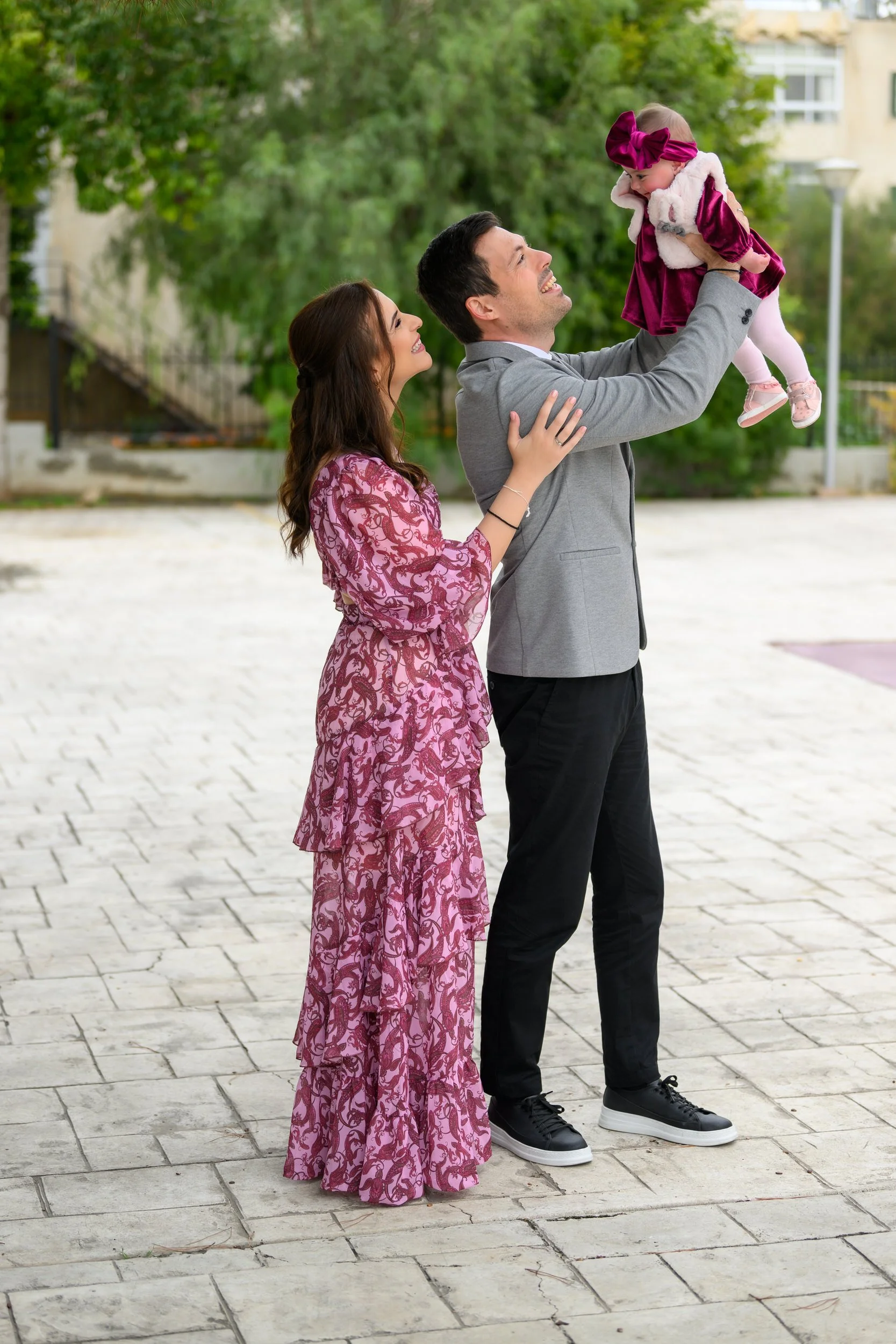 A couple is outdoors, with the man lifting a young girl dressed in pink and purple. The woman stands close, smiling at them, on a paved area with trees and buildings in the background.