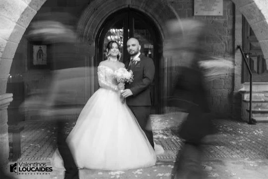 Bride and groom posing for wedding photo in front of a church door, with blurred guests walking past.