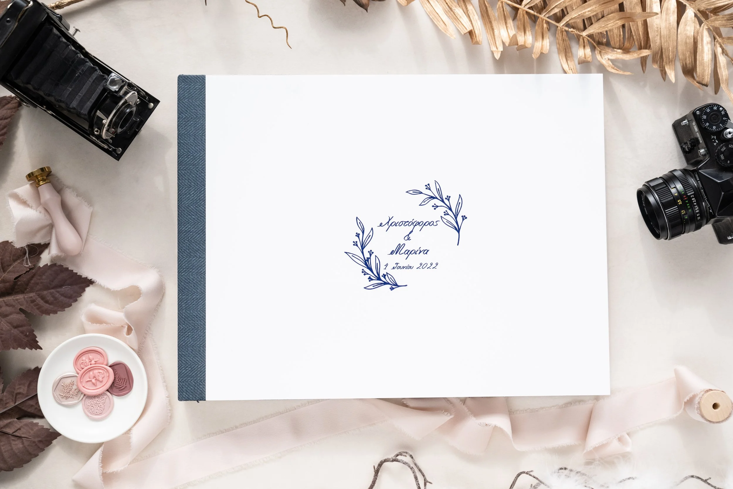 A white wedding album with Greek text and floral design on the cover, surrounded by vintage cameras, ribbon, leaves, buttons, and fabric, on a white surface.