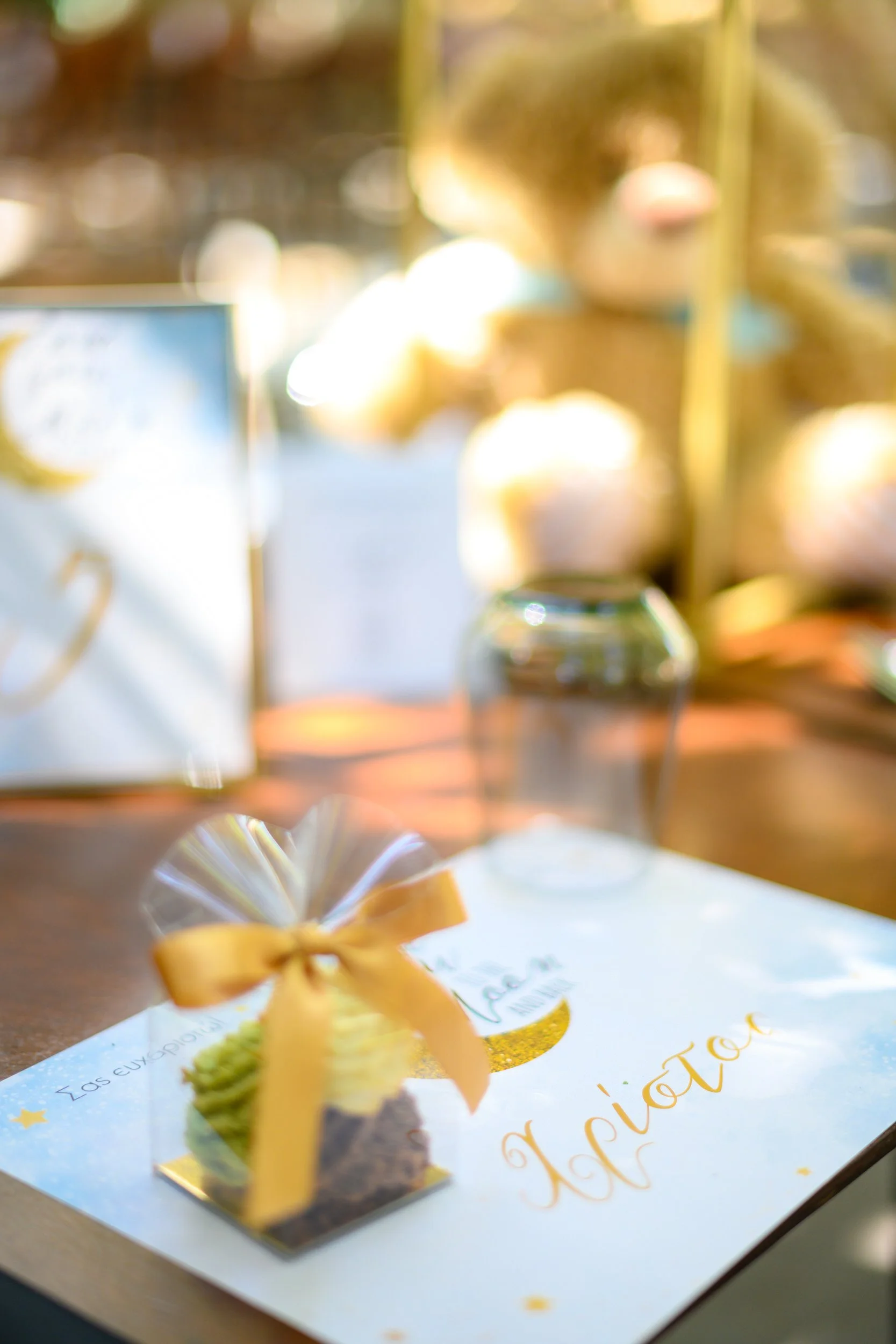 A small gift box with a yellow ribbon on a table, with a card with golden writing and decorations underneath, and blurred balloons and a sign in the background.