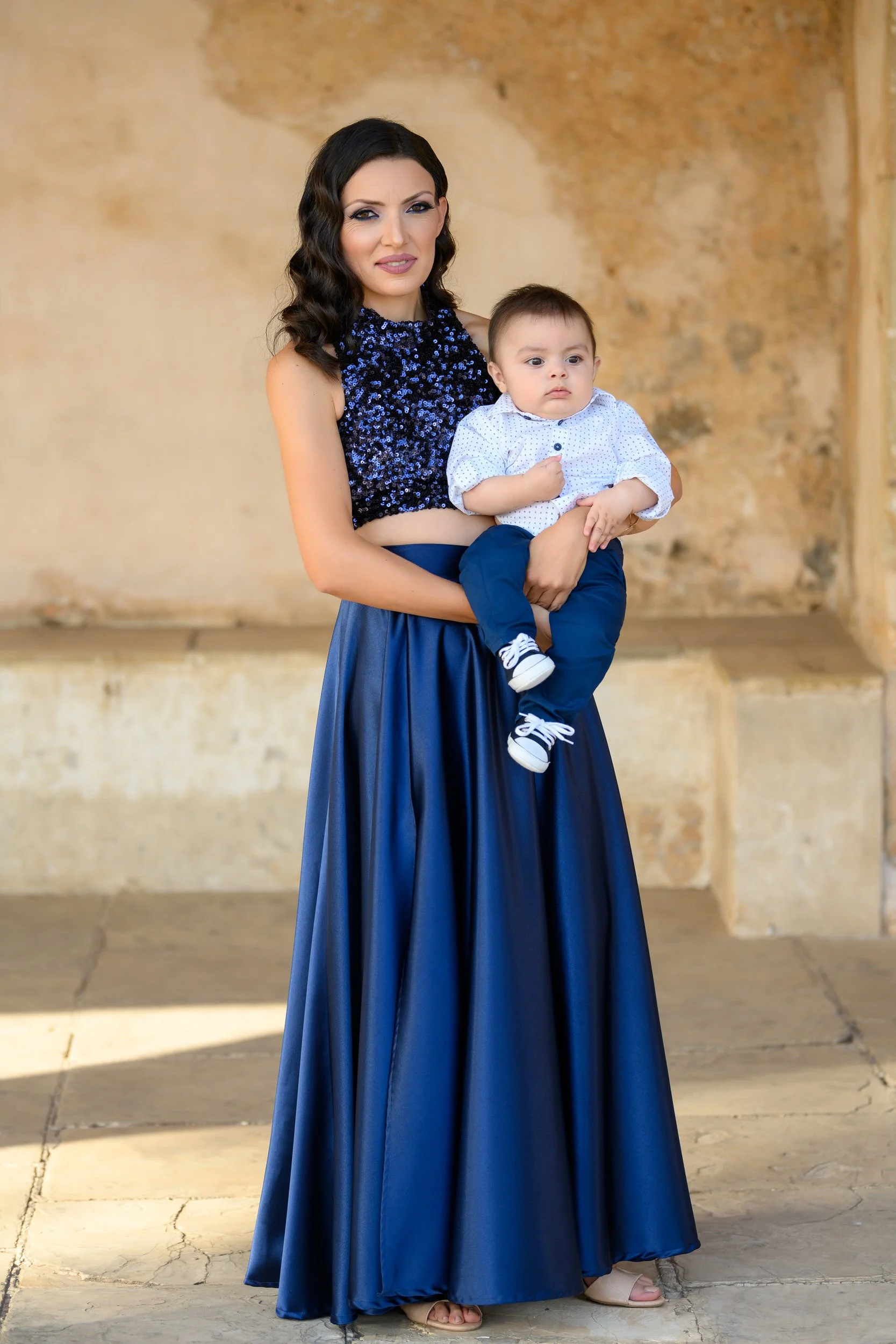 A woman with black wavy hair holding a young boy. She is wearing a sleeveless, dark blue, sequined crop top and a long, flowing blue skirt. The boy has short brown hair and is dressed in a white shirt with small blue polka dots, dark blue pants, and 