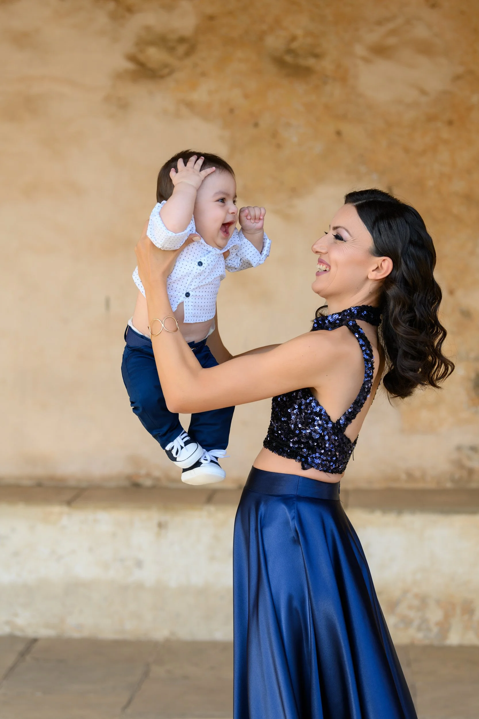 A woman holding a happy toddler boy in the air, both smiling and looking at each other indoors with a beige textured wall in the background.