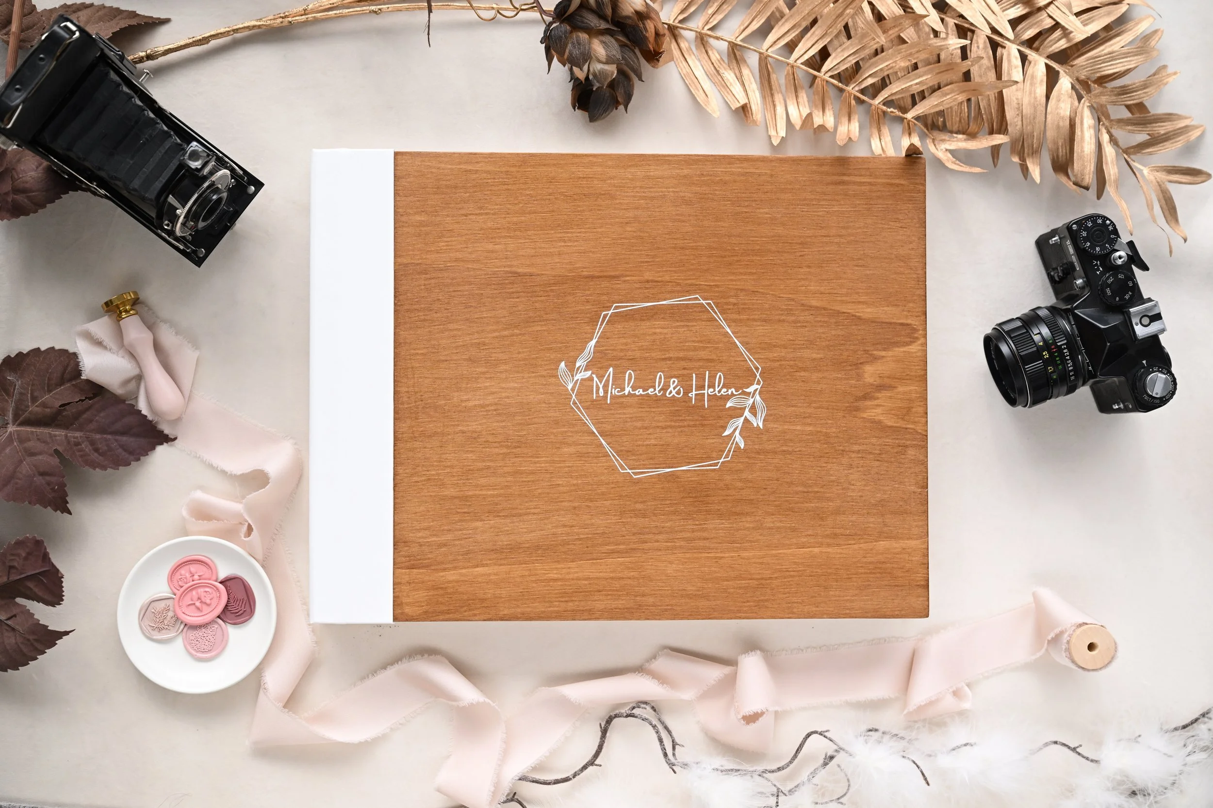 A wooden wedding album cover with the names Michael and Helen inscribed in a decorative style, surrounded by photography equipment, dried plants, and decorative items on a table.