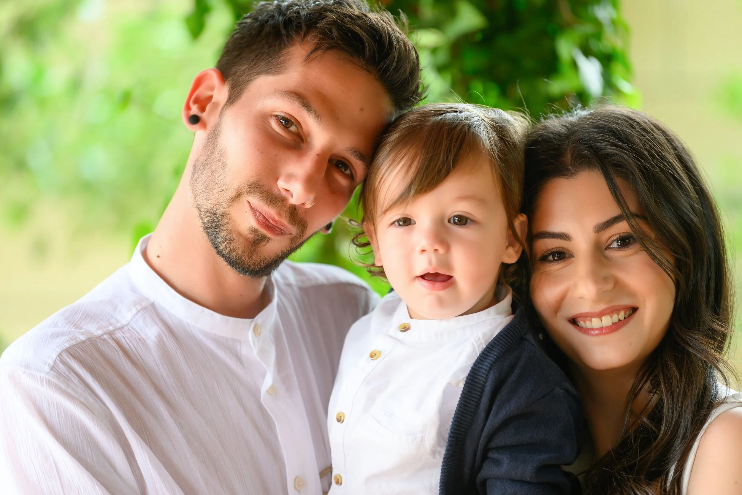 A happy family of three, a man, woman, and child, smiling and close together outdoors with greenery in the background.