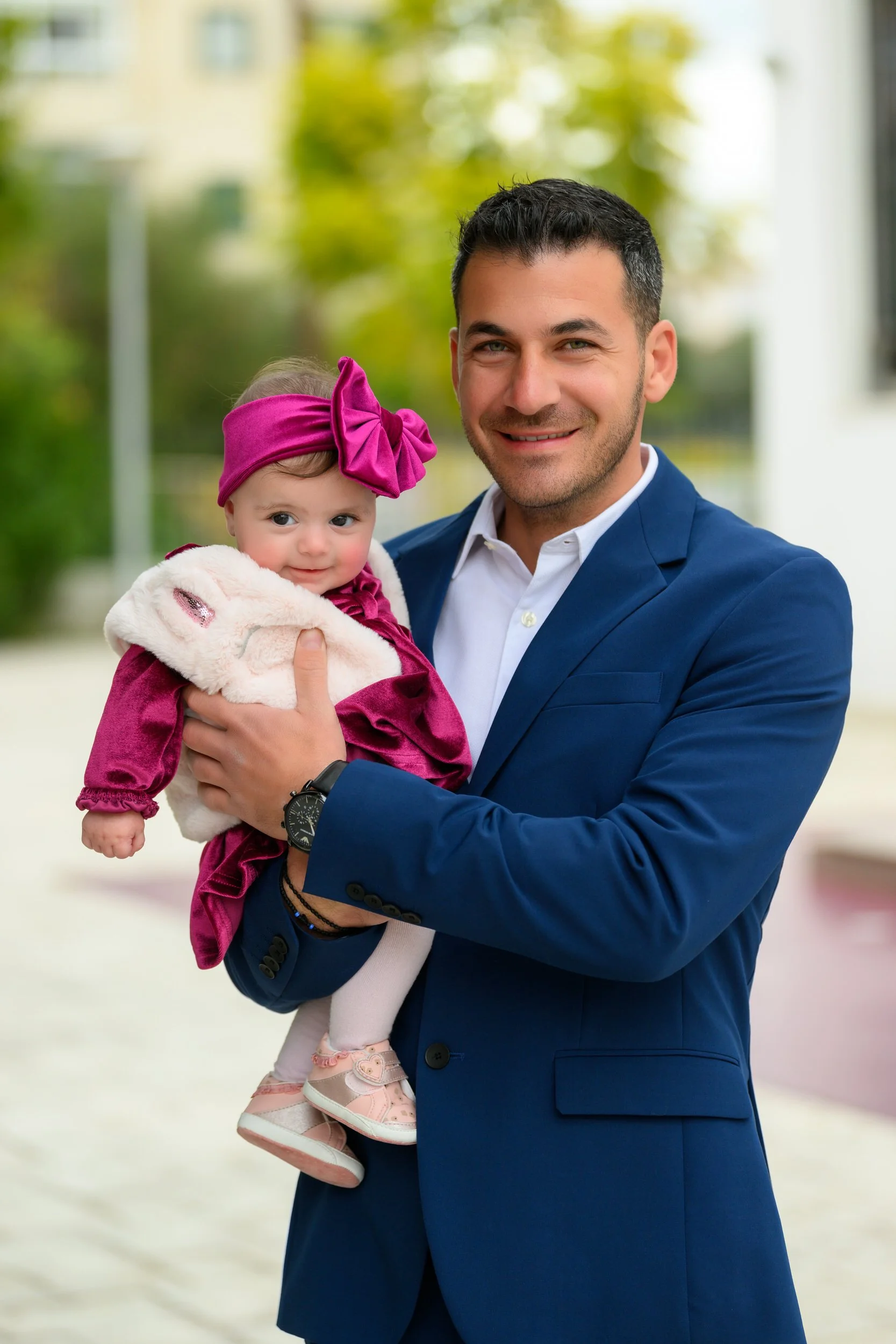 A man in a blue suit holding a young girl in a pink dress and headband outdoors with blurred trees and buildings in the background.