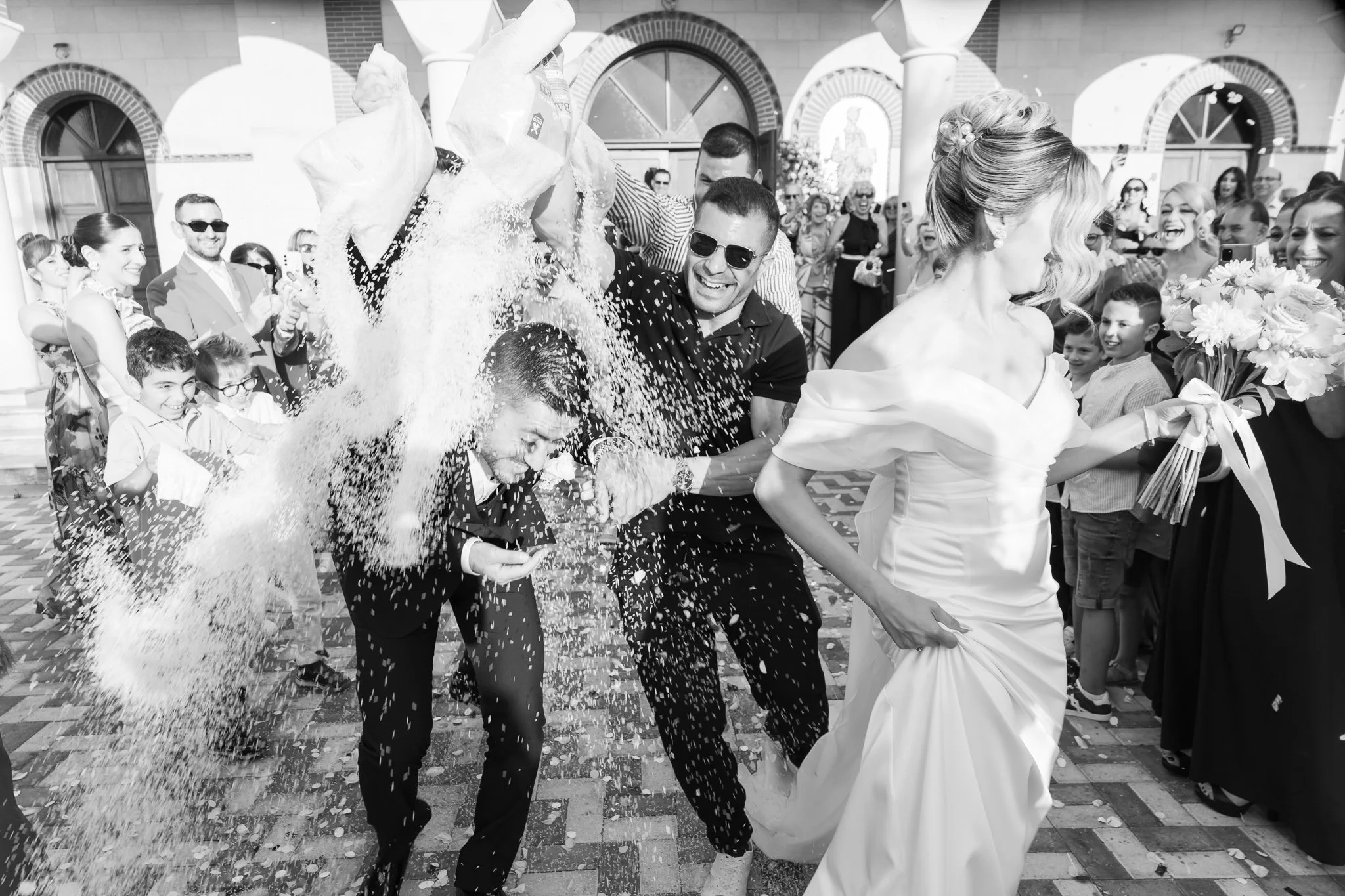 Bride in a white dress and groom in a suit, surrounded by smiling guests, some children, at a wedding celebration. The groom is pouring rice or confetti over the bride as part of a wedding tradition.