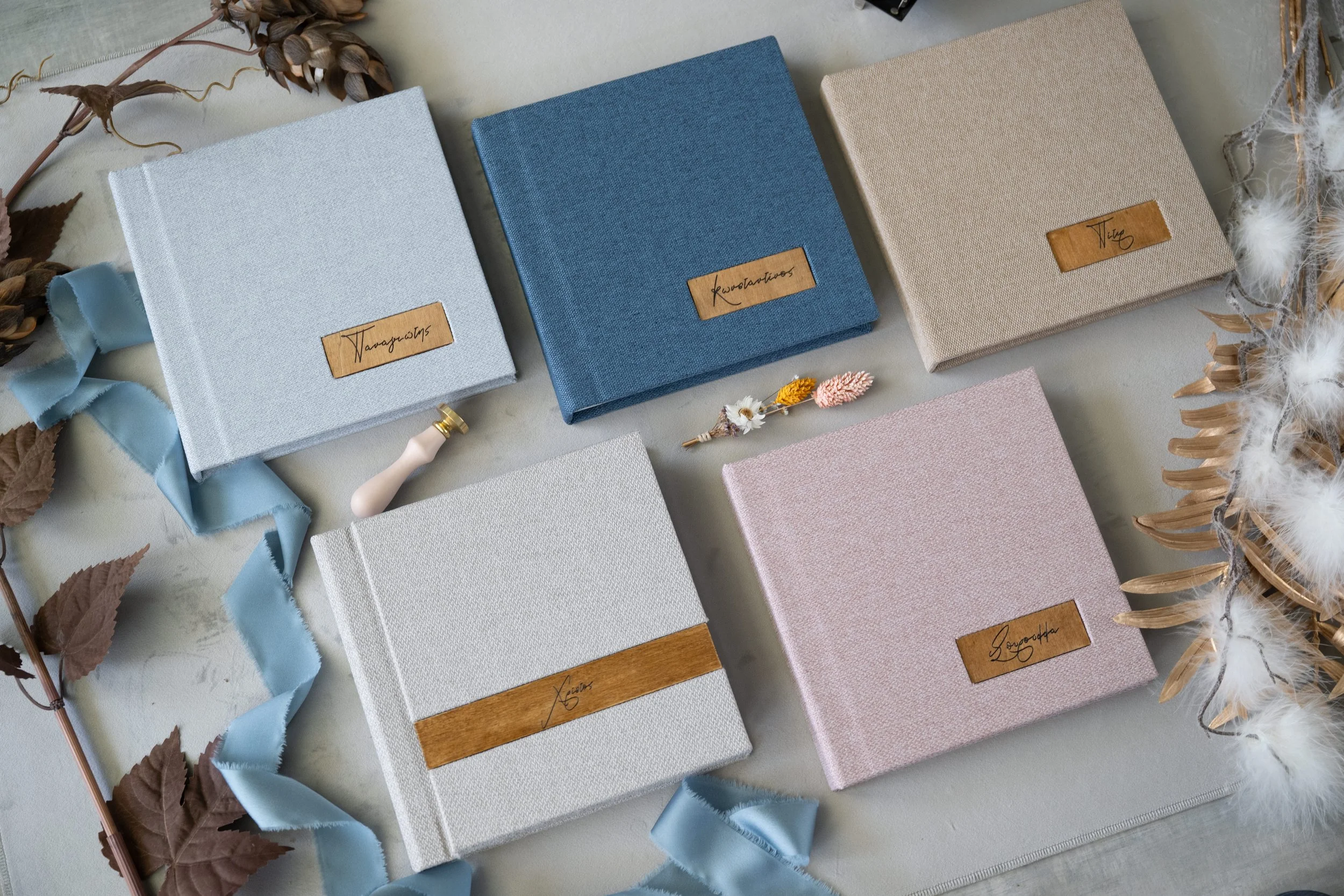 Six fabric-covered lay flat albums with wooden labels on a light surface, surrounded by dried leaves, a blue ribbon, and white feathers.