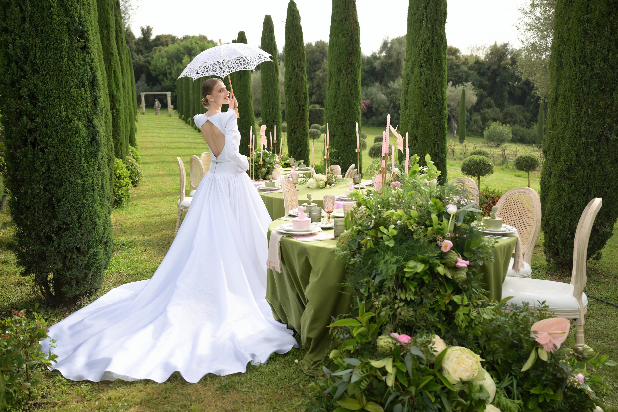 A bride in a white wedding gown holding an umbrella, standing next to a decorated outdoor dining table with greenery and flowers in a garden setting.