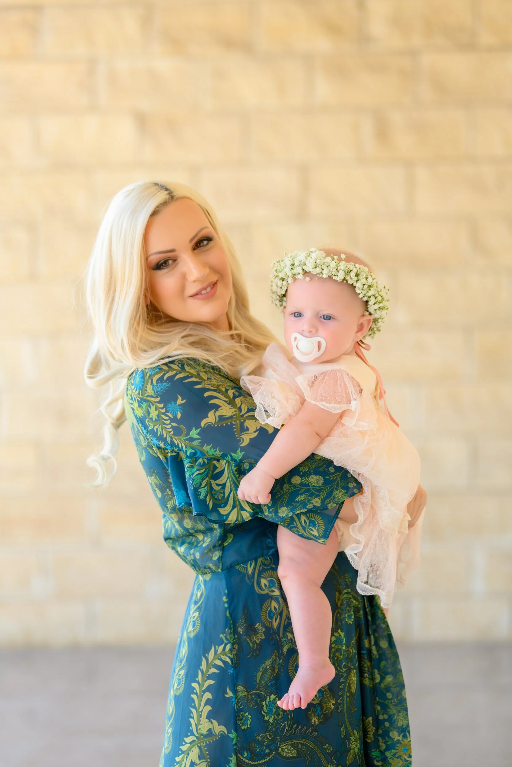 A woman with blonde hair holding a baby girl with a floral crown against a light blurred background.