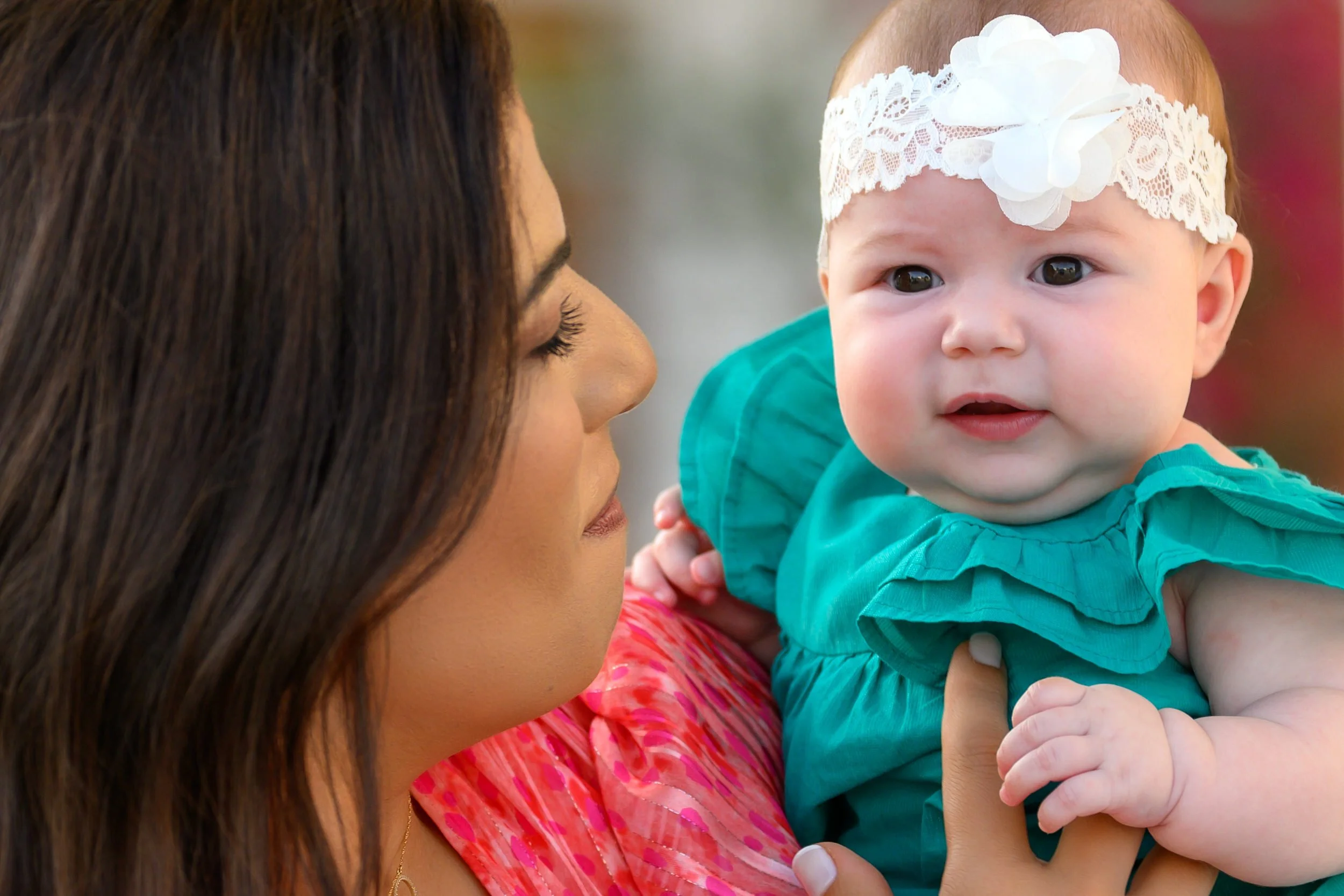 A woman holding a baby girl outdoors, with the woman looking at the baby who is wearing a white lace headband and a teal dress.