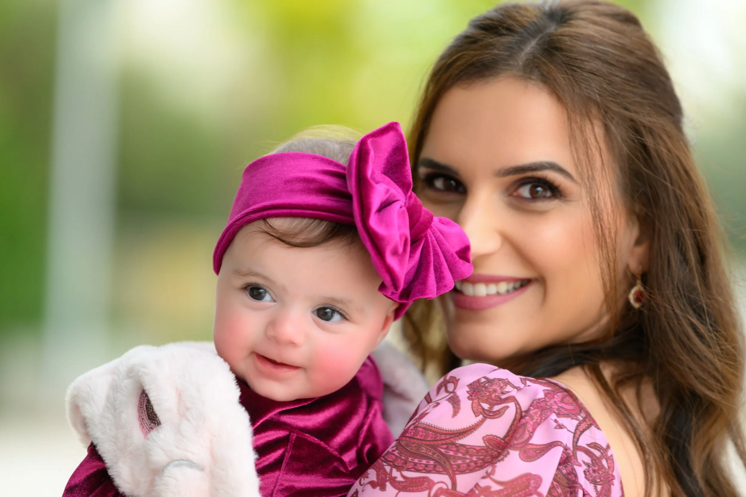 A woman smiling while holding a young girl dressed in pink and purple, with a pink headband and bow, outdoors with a blurred green background.