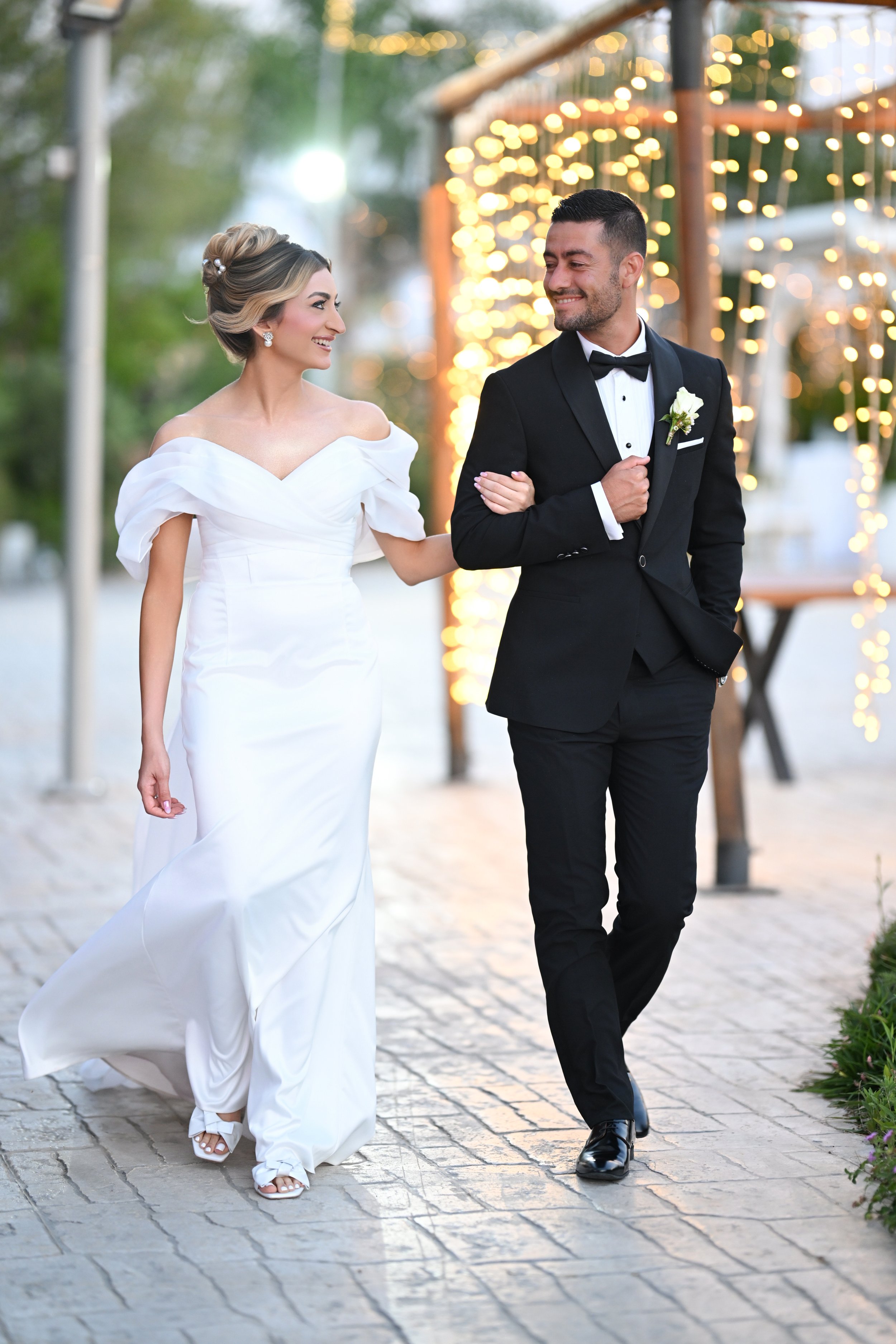 A bride and groom walking outdoors during their wedding celebration, with string lights in the background, the bride in a white gown and the groom in a black tuxedo.