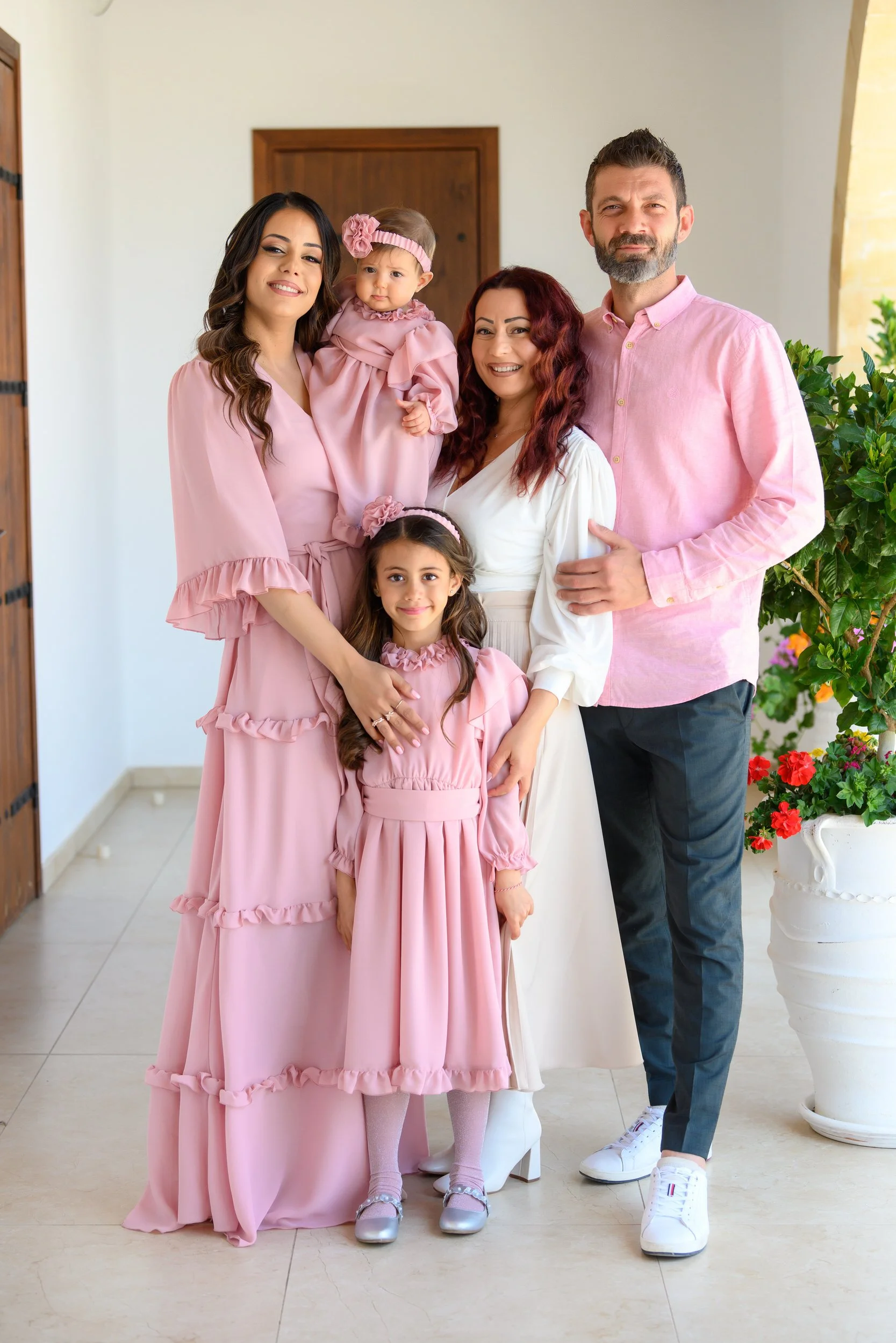 A family of six posing together inside, with three adult women, a man, and two young girls, all dressed in pink and white dresses, smiling happily.