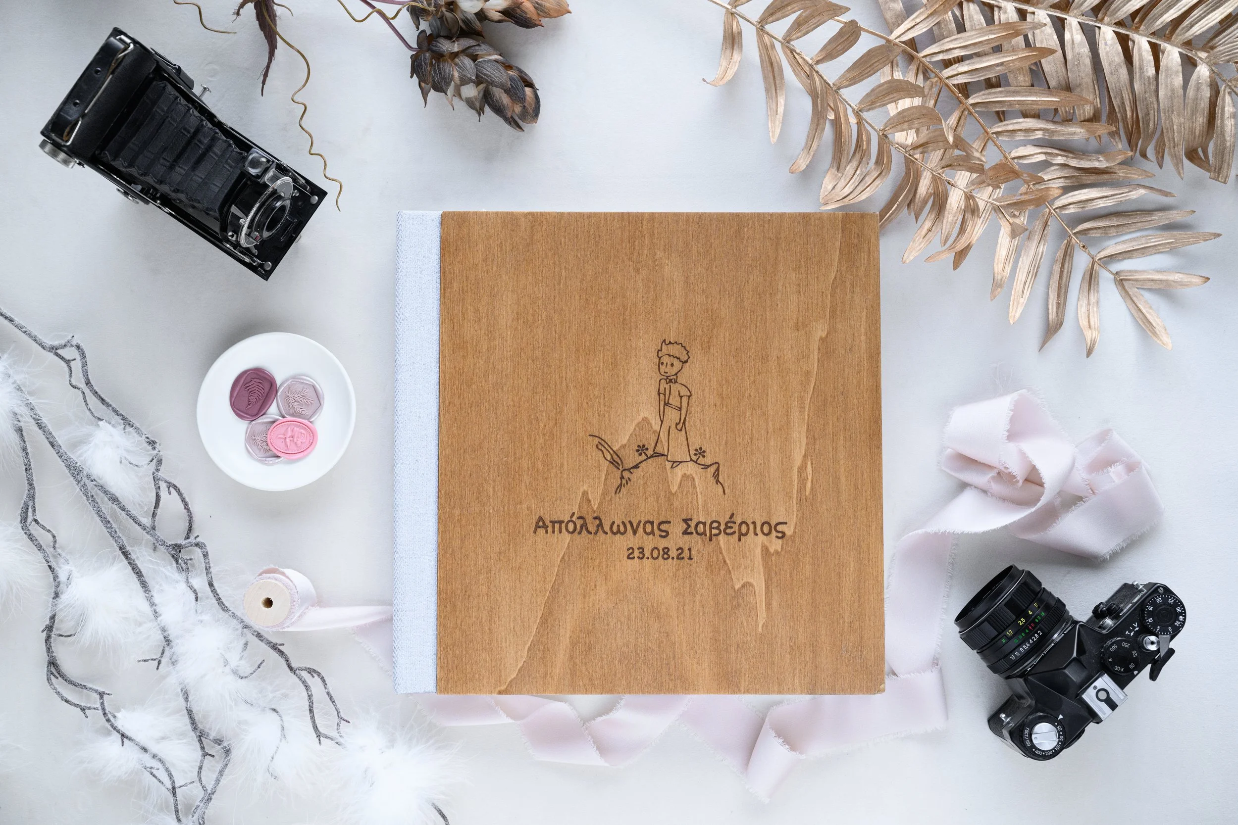 A wooden lay flat album with an engraved illustration of a person and Greek text, surrounded by vintage cameras, dried plants, silk ribbon, decorative buttons, and feathers on a white surface.