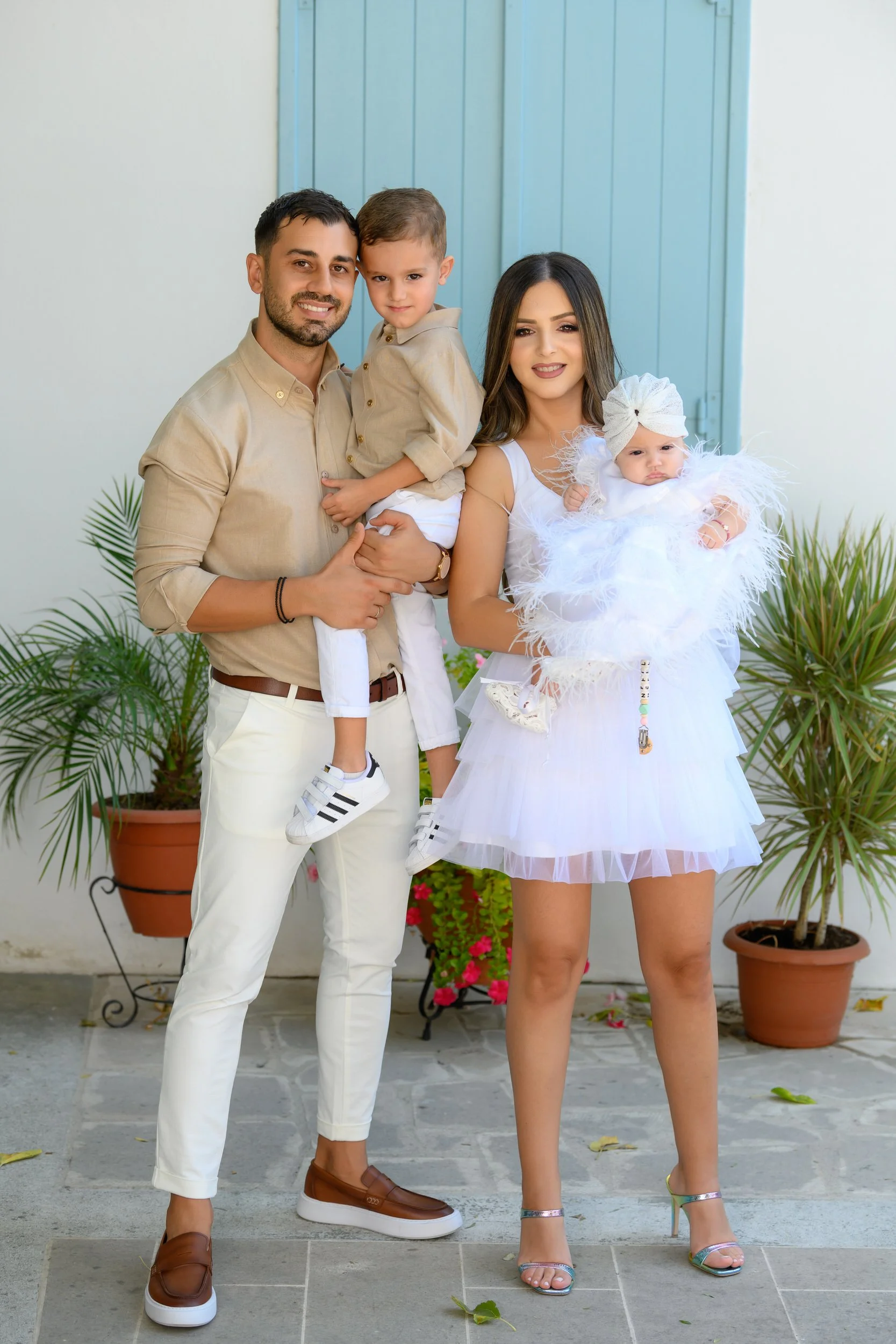 A family of four standing outside in front of potted plants and a light blue door. The father is holding a young boy, and the mother is holding a baby girl dressed in white. The family is dressed in coordinated neutral and white outfits.