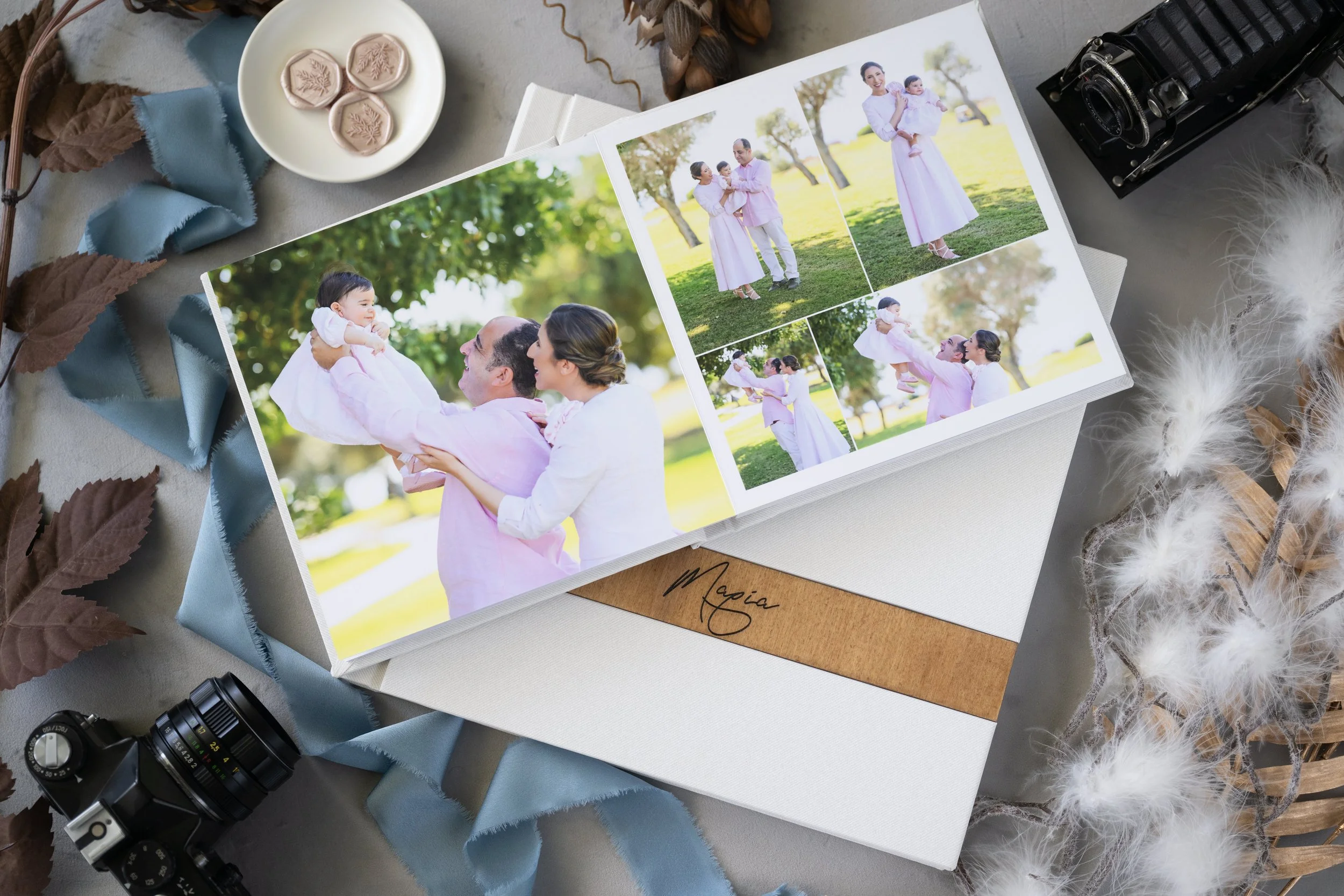 An open photo album displaying pictures of a family with a baby, surrounded by decor including a vintage camera, leaves, ribbon, and fluffy white feathers on a surface.
