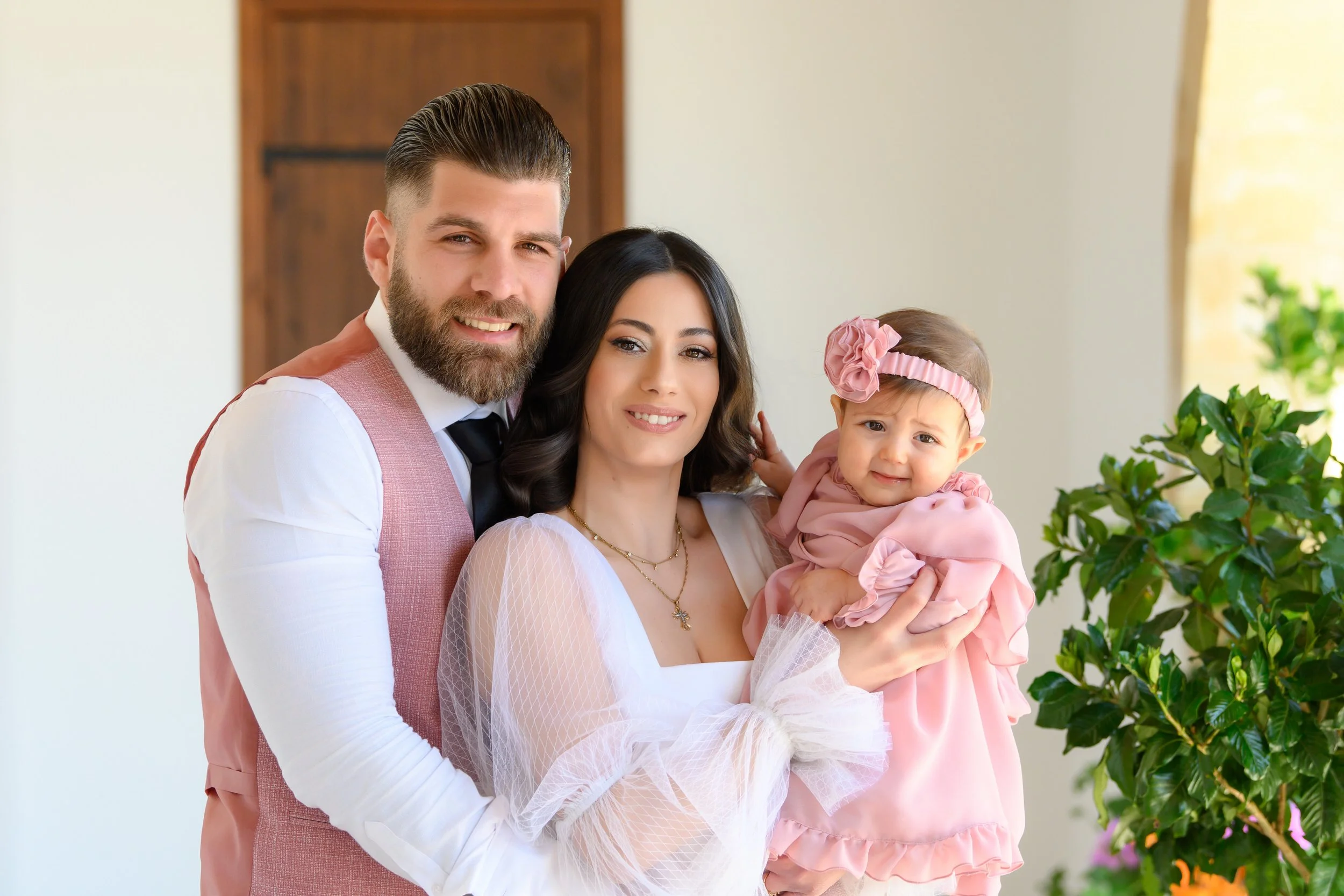 A happy family of three with a father, mother, and young daughter, dressed in semi-formal attire, standing indoors near some greenery and smiling at the camera.