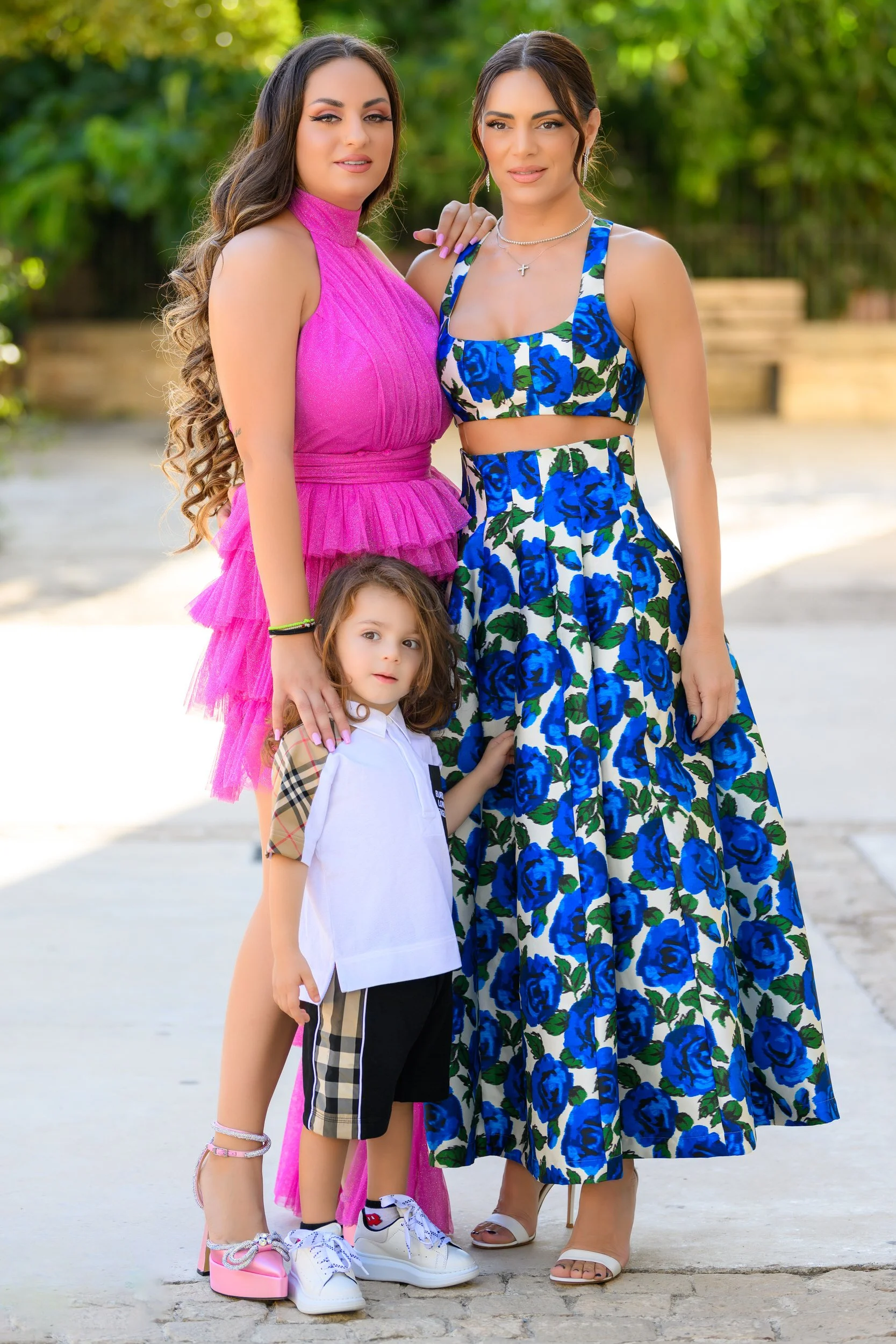 Two women and a young girl standing outdoors on a sunny day. One woman is wearing a long blue and white floral dress, and the other is in a bright pink dress. The young girl is in front, wearing a white hoodie with Burberry patterns on the sleeves an