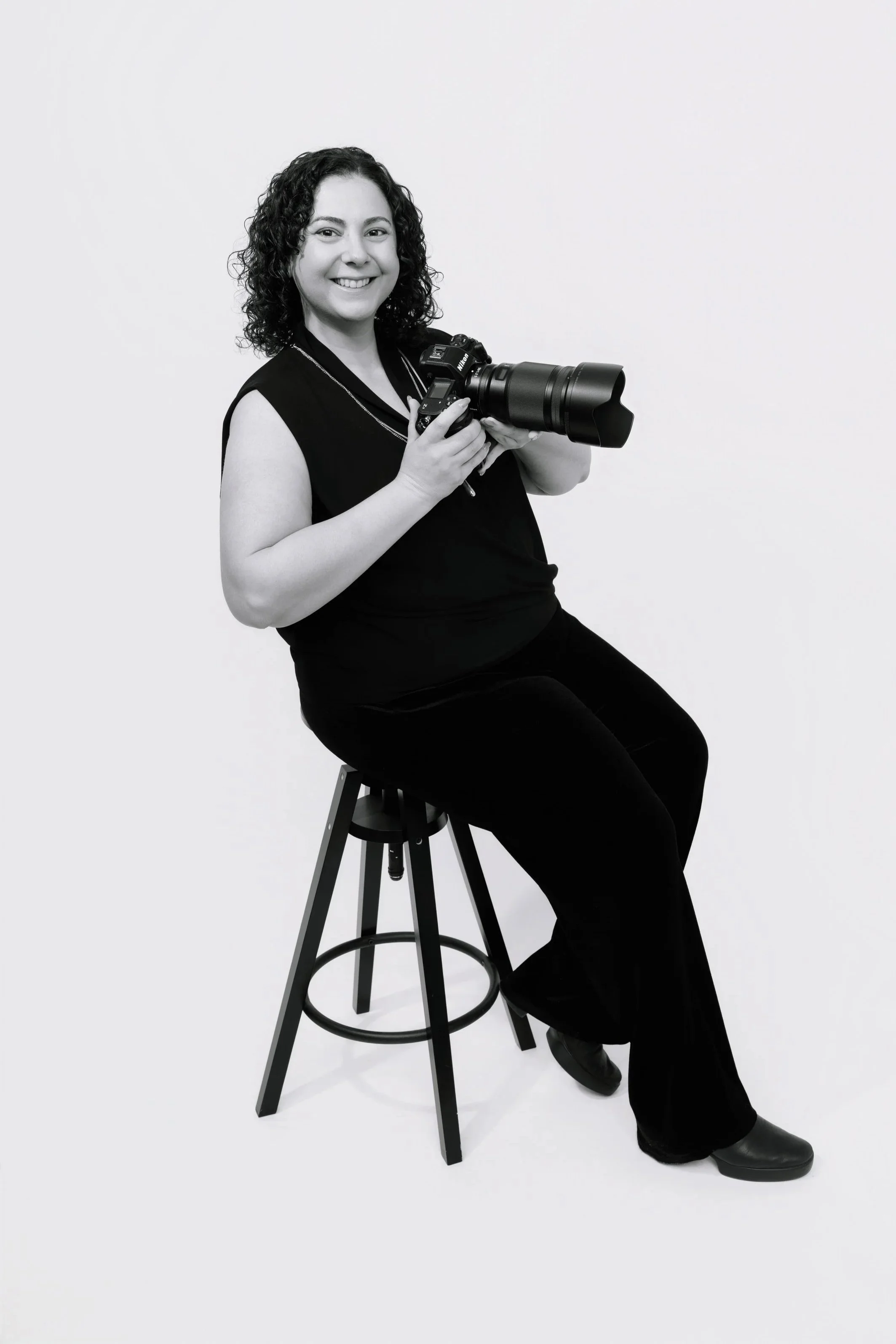 A woman with curly hair sitting on a stool, holding a professional camera with a large lens, smiling at the camera against a plain white background.