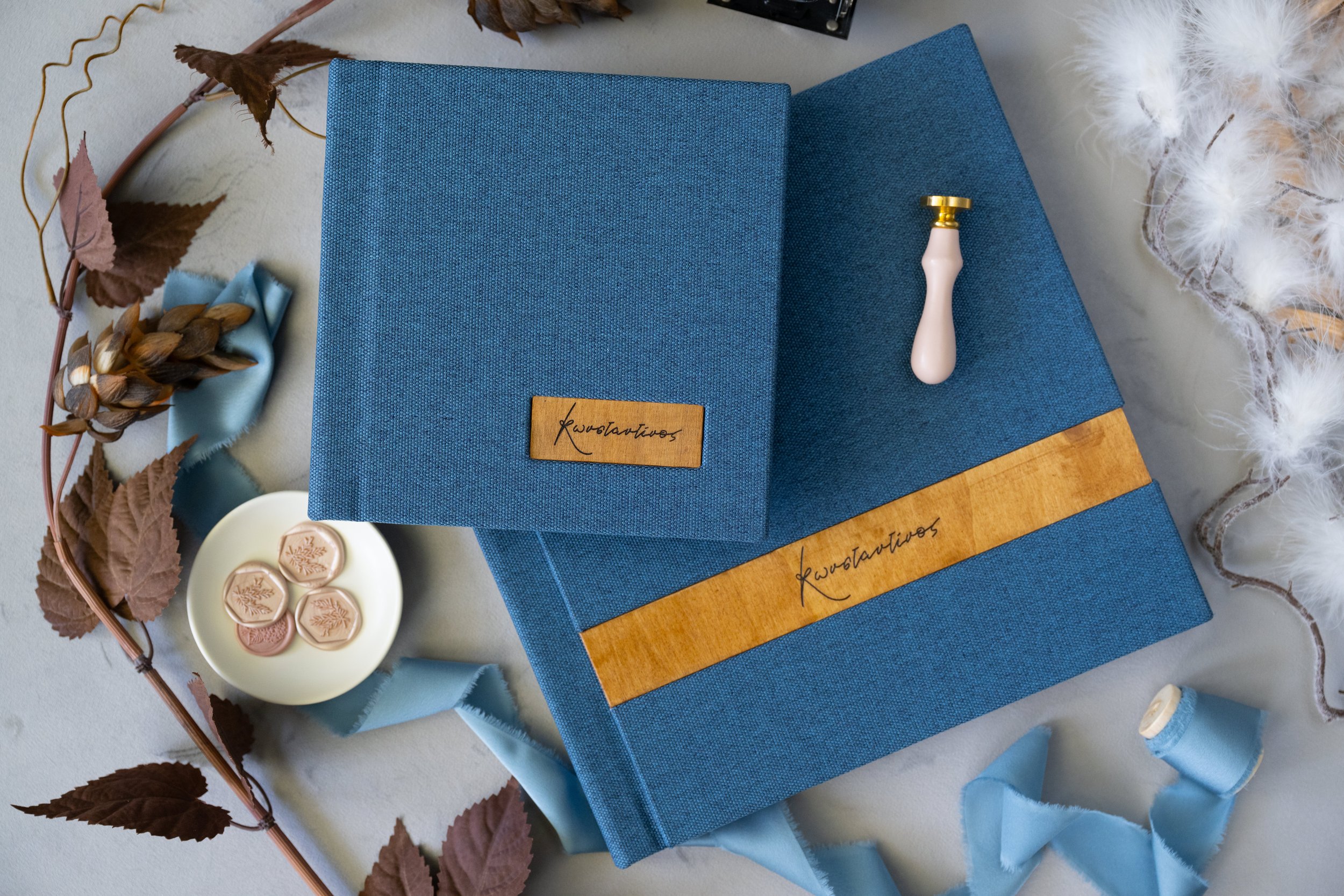 Blue wedding guest book with a wooden band labeled "Kwestantivos", a wax seal stamp with a pink handle, and a white plate of wax seals, surrounded by dried leaves, blue satin ribbon, and white feathered decorations.