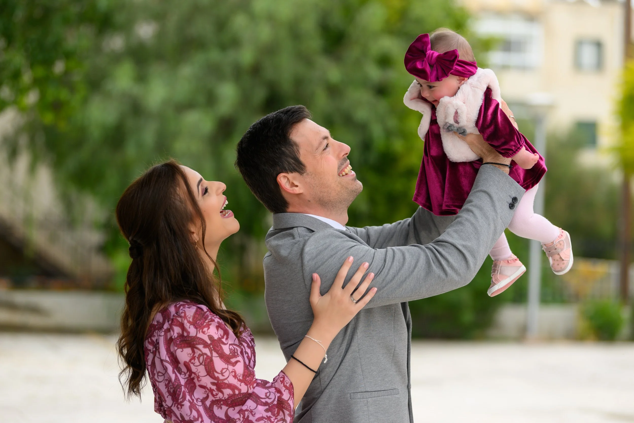A man in a gray suit is lifting a young girl in a maroon dress and matching headband with a large bow, while a woman with long brown hair in a pink patterned dress looks on happily in an outdoor park setting.