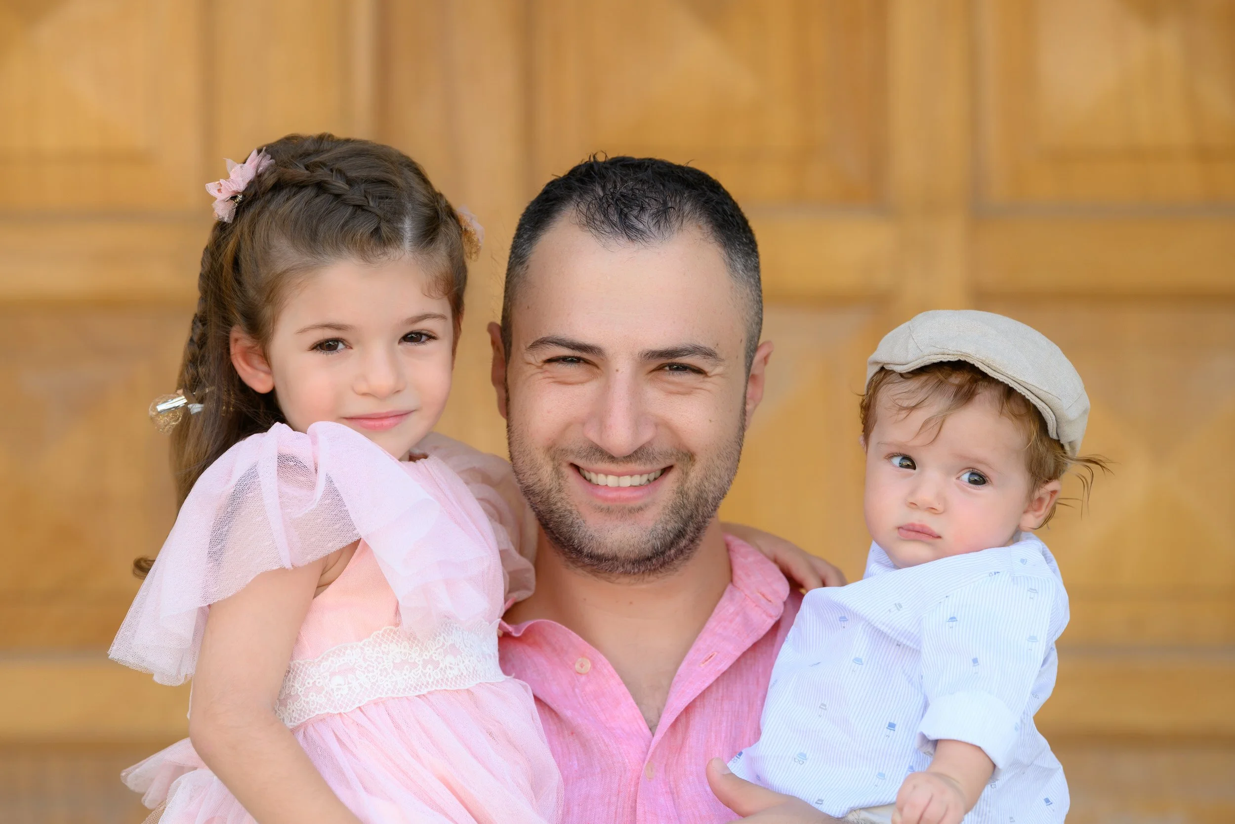 A man smiling and holding a young girl in pink dress with pigtails, and a young boy in a light blue shirt and beige cap, in front of a wooden background.