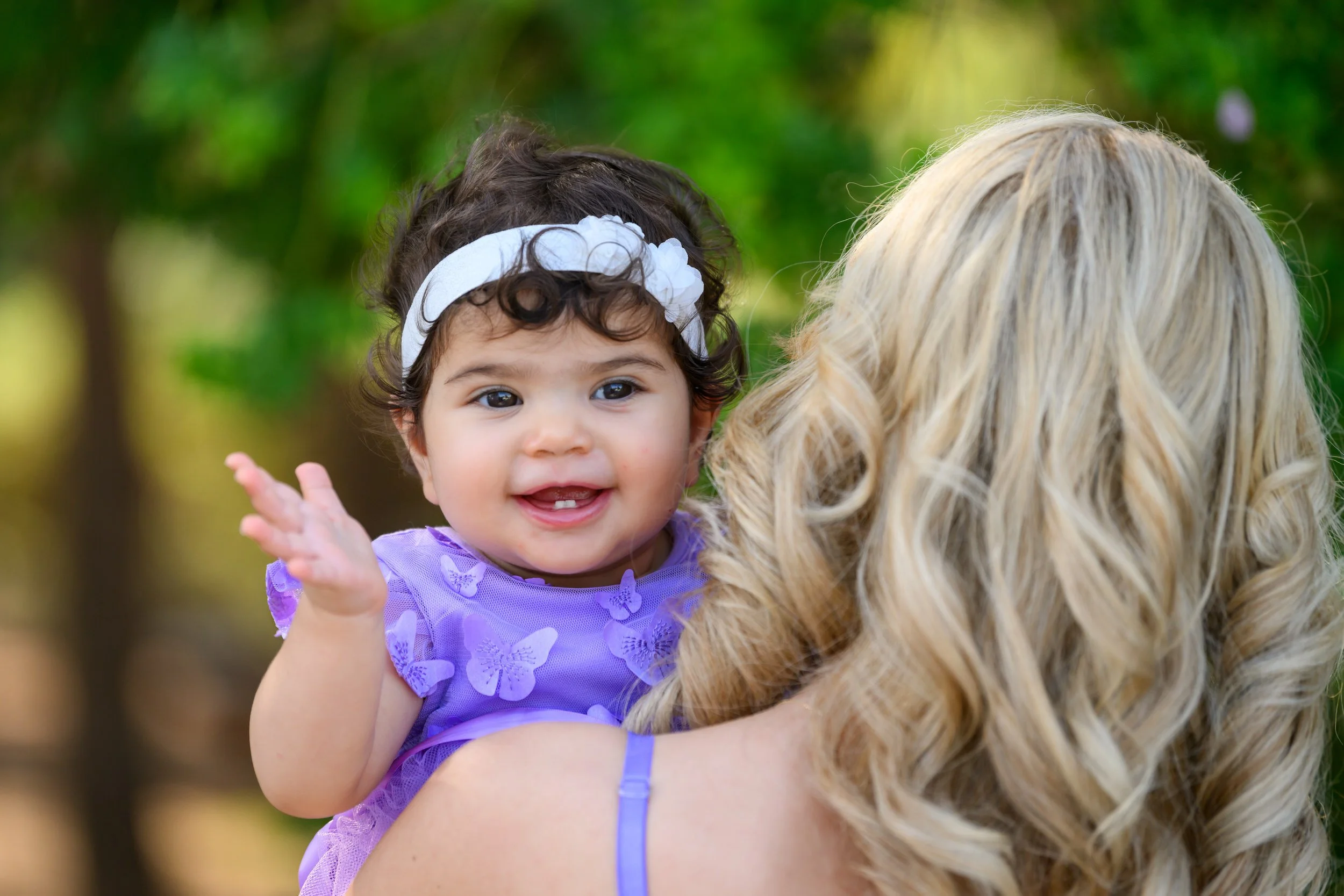 A smiling young girl with dark curly hair, wearing a white headband and a purple dress with butterfly patterns, is being held by a woman with long blonde curly hair in an outdoor setting with green trees in the background.