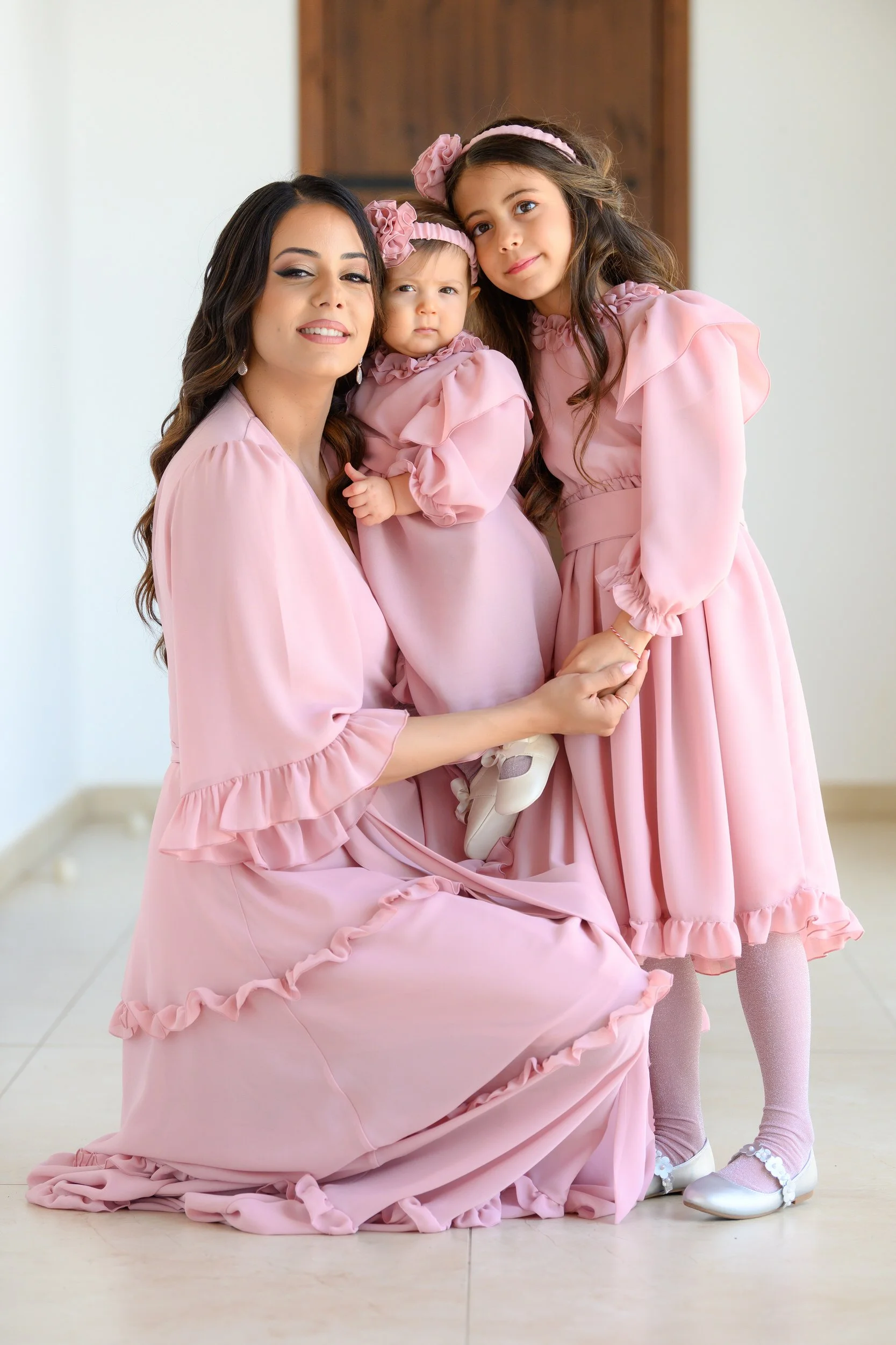 A woman and two young girls dressed in matching pink dresses pose together indoors, with the woman kneeling and holding the younger girl, while the older girl stands beside them.