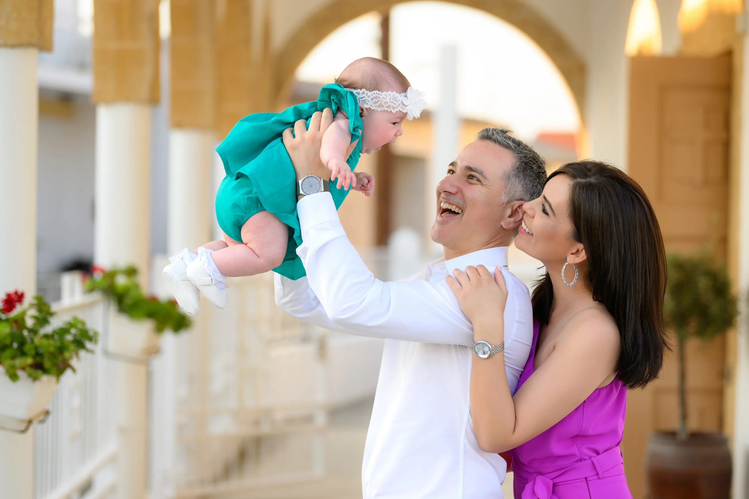 Happy family outdoors, father lifting baby girl, mother smiling, bright background.