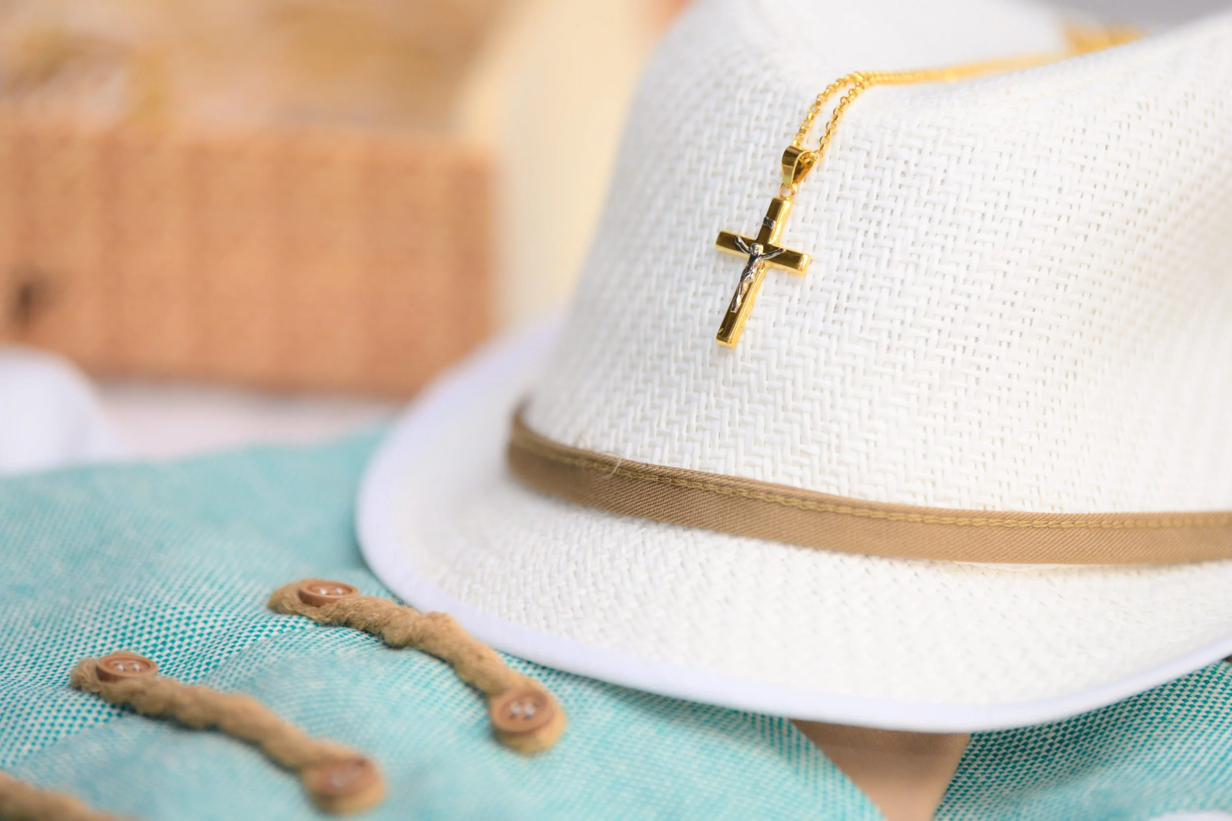 A white hat with a beige band and gold chain, displaying a gold and silver cross necklace, resting on a light blue fabric with brown buttons and beige trim, with a blurred background.