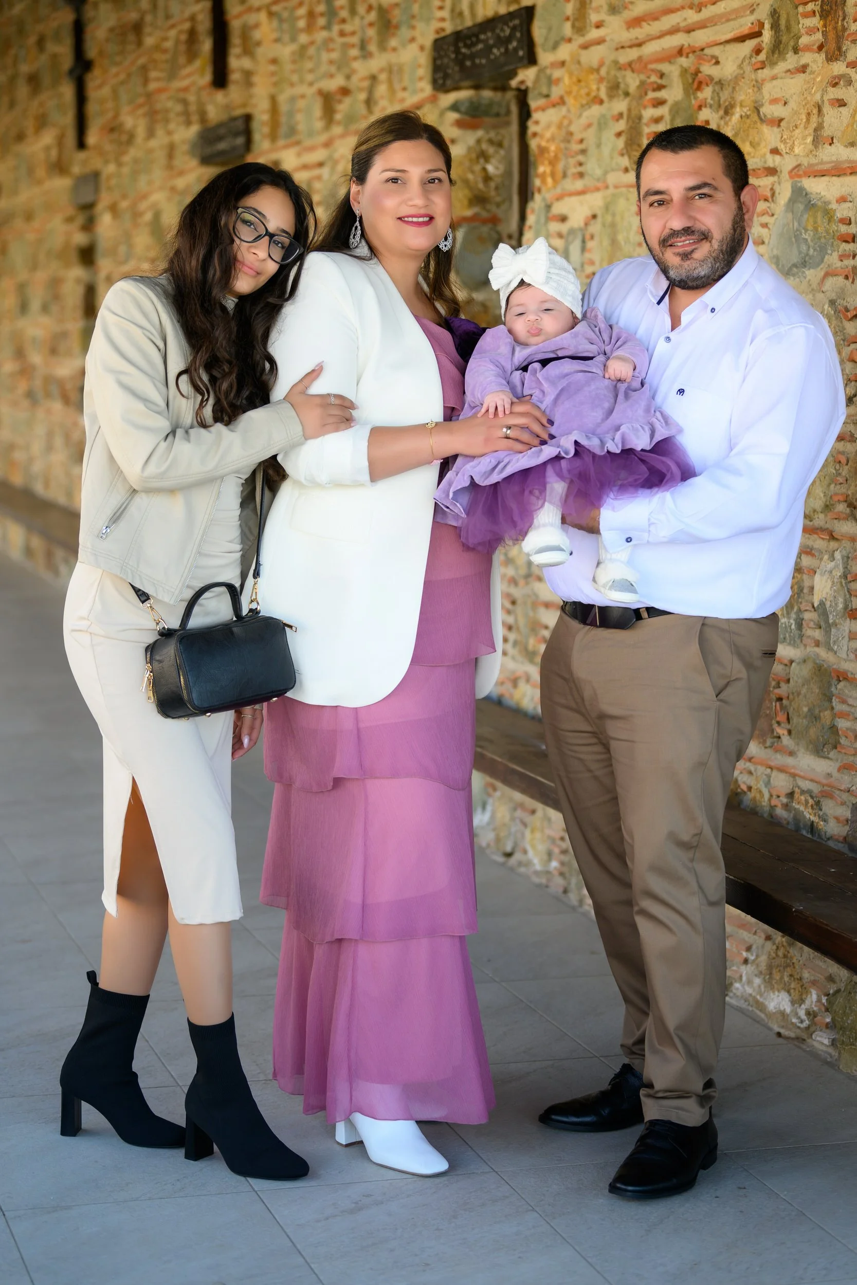 A family of four standing against a stone wall, including a young woman with glasses in a beige jacket and white skirt, a woman in a white blazer and pink dress, a man in a white shirt and beige pants holding a baby girl dressed in purple with a whit