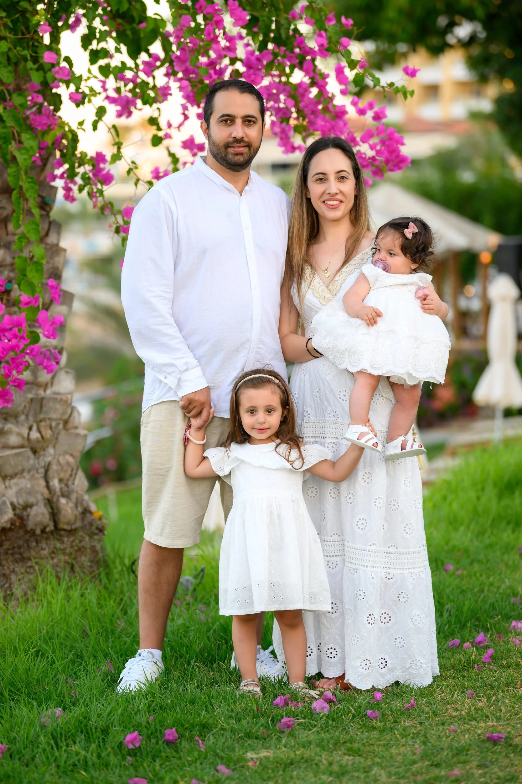 Family of four, two adults and two young girls, posing outdoors under blooming pink bougainvillea. The woman holds a young girl in a white dress, the man stands beside her, and the other girl in a white dress stands in front, holding the woman's hand