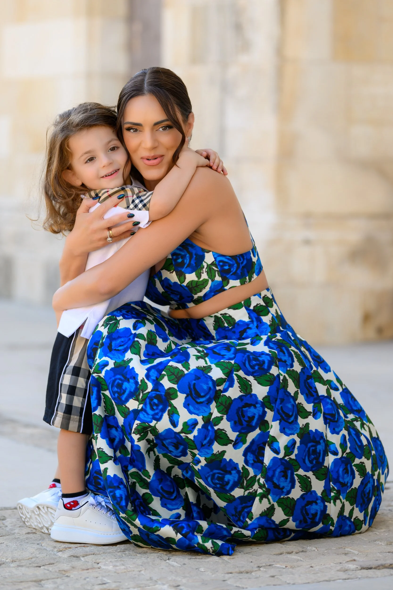 A woman in a blue floral dress hugging a young girl with curly hair, both smiling, outdoors with stone walls in the background.