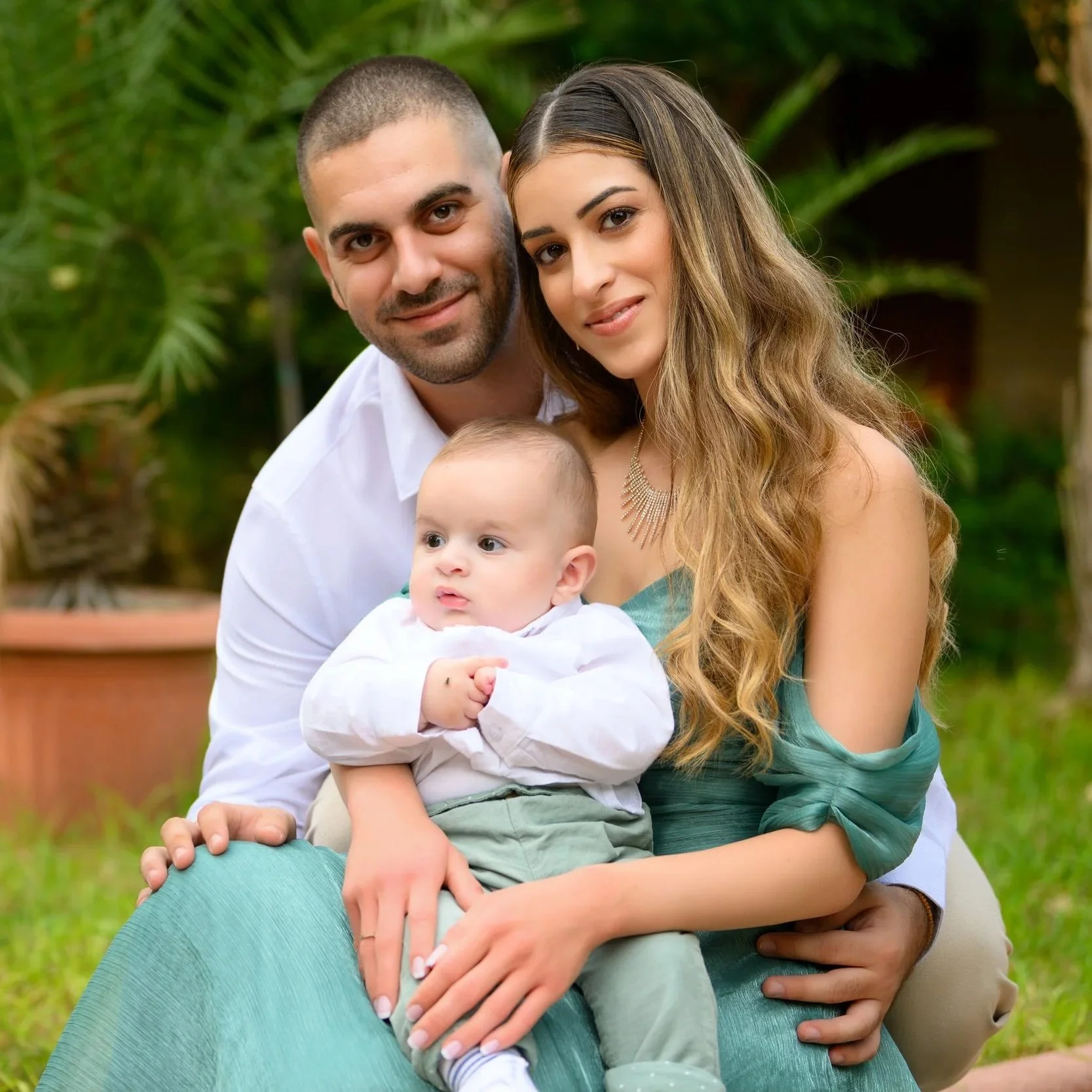 A happy family of three sitting outdoors with green plants in the background. The father has short dark hair and a beard, wearing a white shirt. The mother has long, wavy, light brown hair and is wearing a teal dress with open shoulders. The young child, sitting on the mother’s lap, has light brown hair, a white shirt, and beige pants. They are all smiling gently.