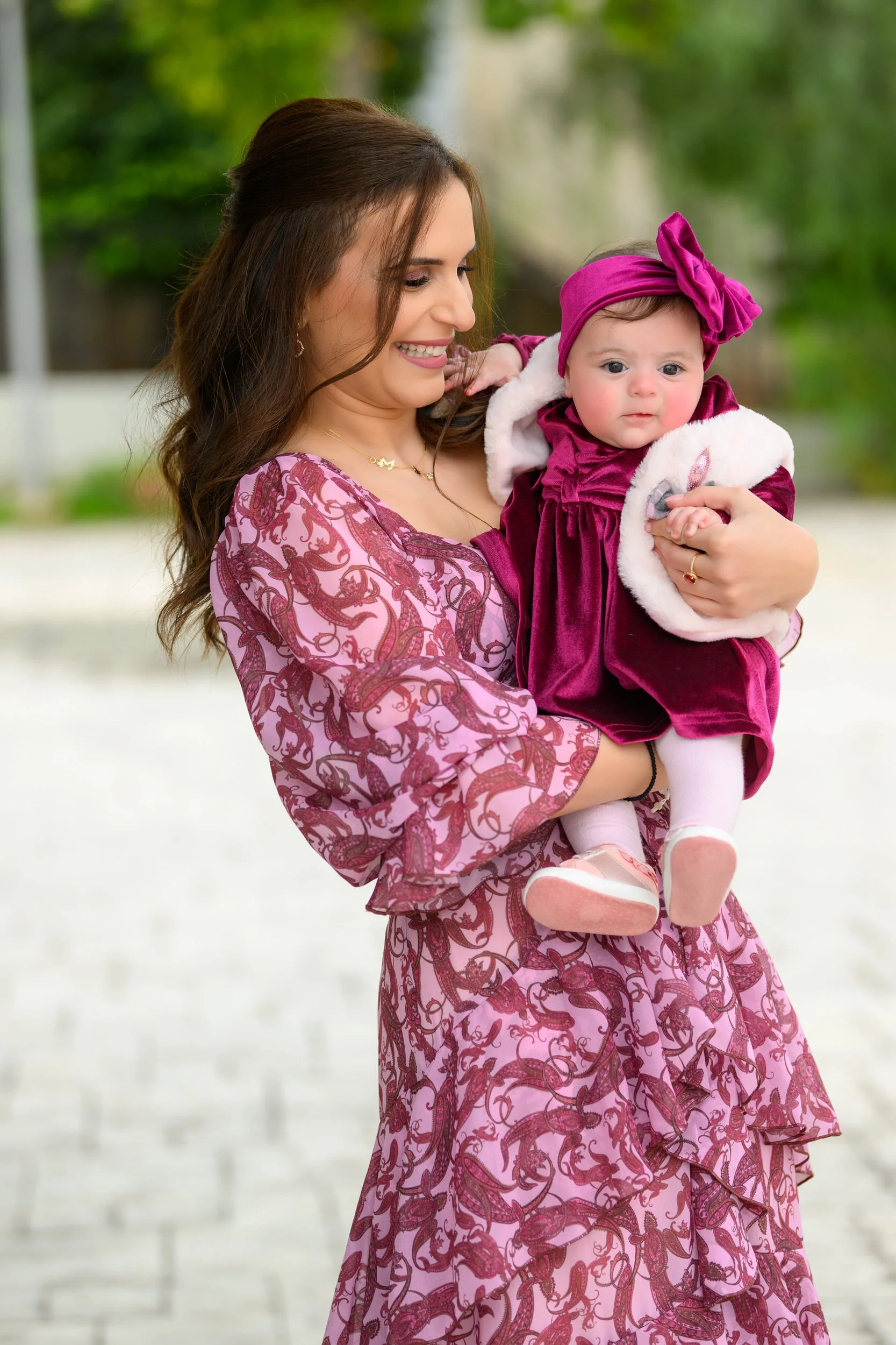 A woman with brown hair wearing a pink and red patterned dress holds a young girl dressed in a velvet burgundy dress with white fur trim and a matching headband, outside in a park on a bright day.