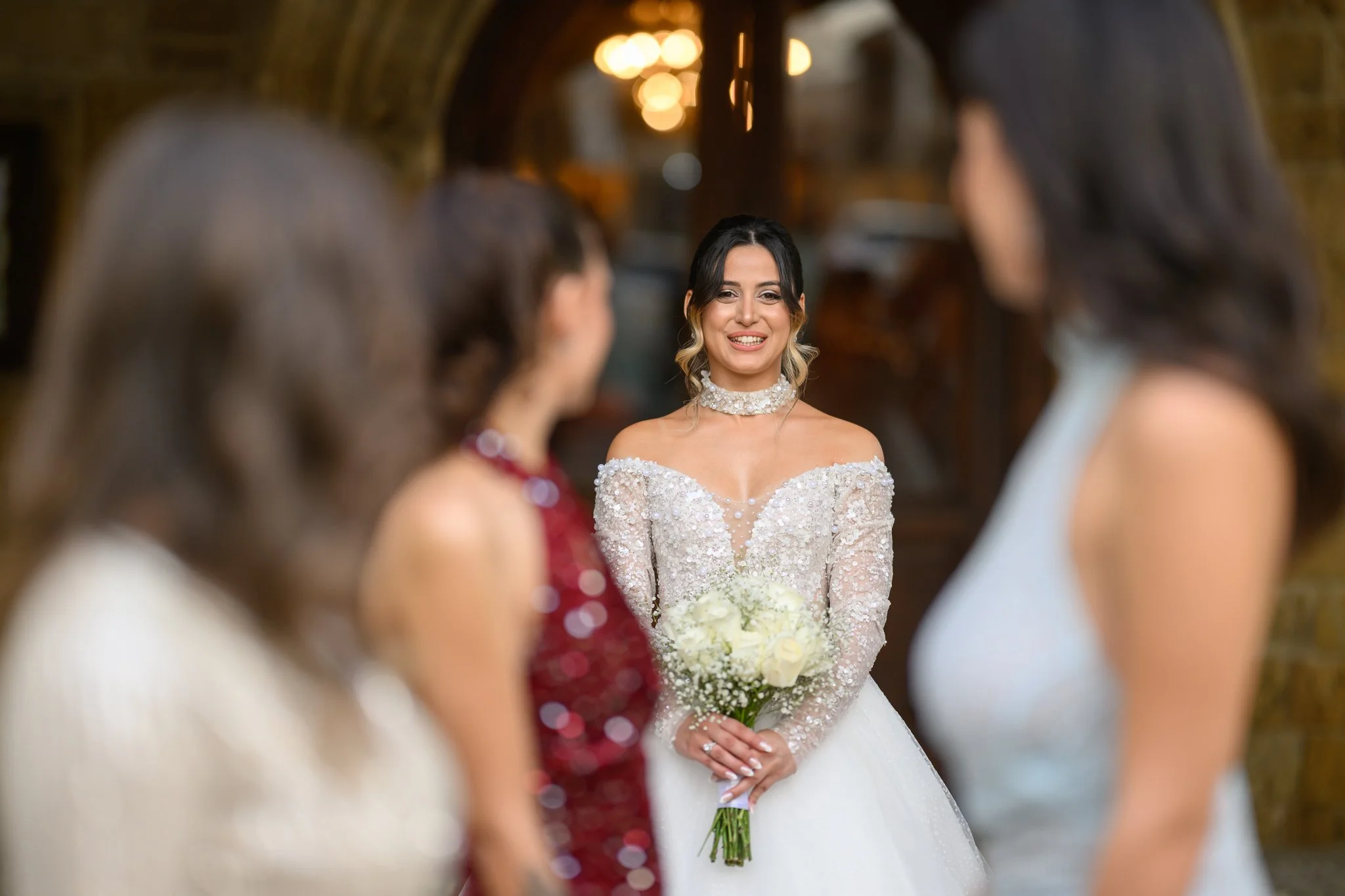 A bride in a wedding gown holding a bouquet of white roses, smiling at three women in colorful dresses at a wedding reception.