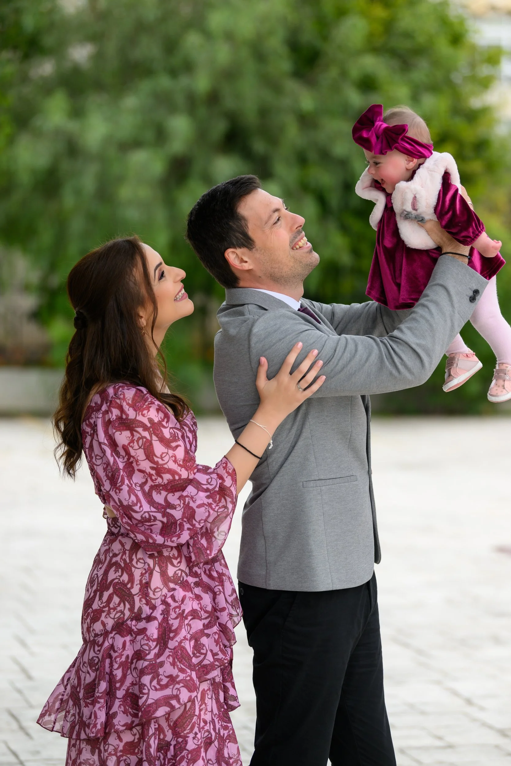 A man lifting a toddler girl in the air while a woman stands beside them, all smiling and looking at each other in an outdoor setting with green trees in the background.