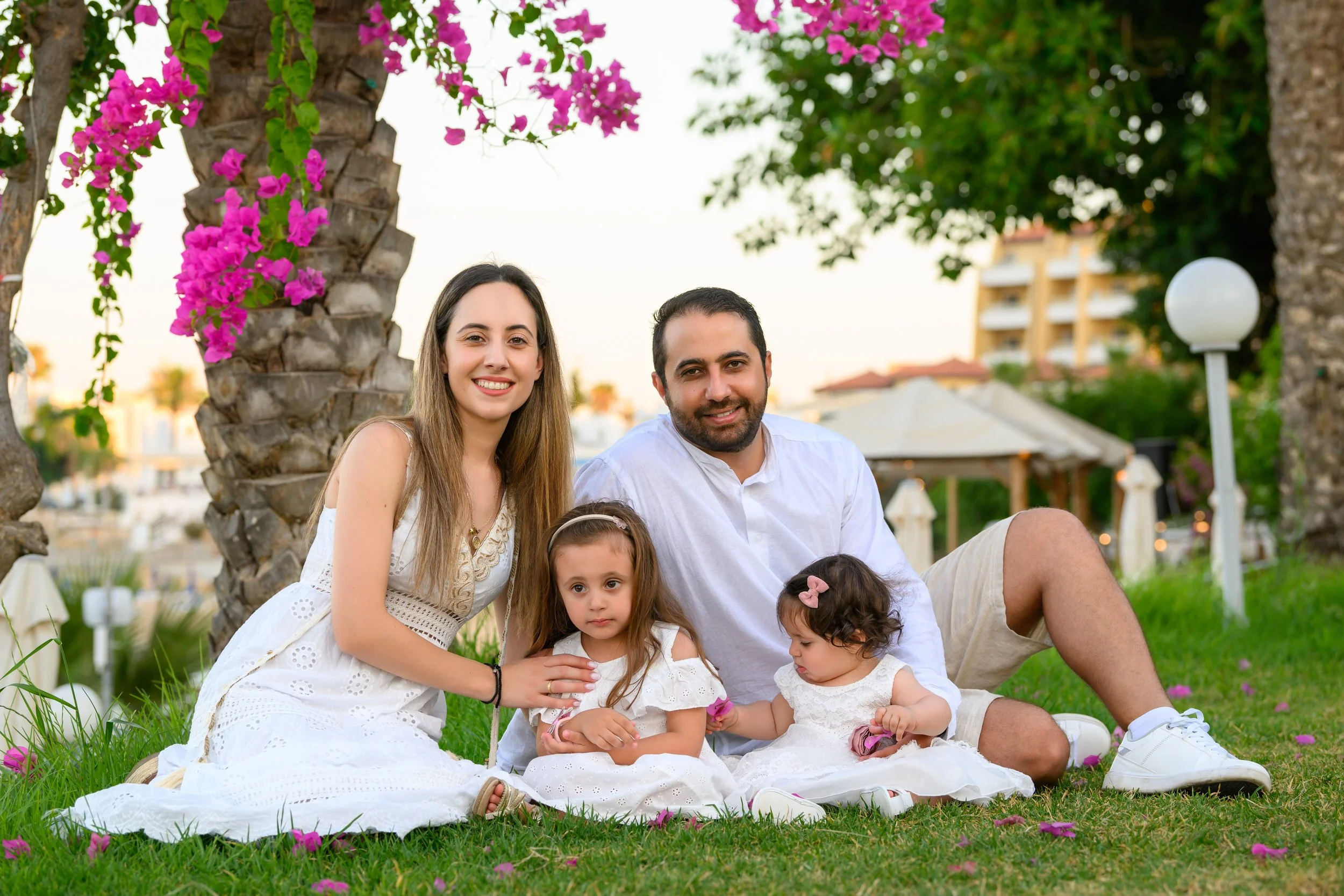 A family of four sitting on the grass in a park or garden with flowering trees and a gazebo in the background. The family consists of a woman, a man, and two young girls, all smiling and dressed in white.