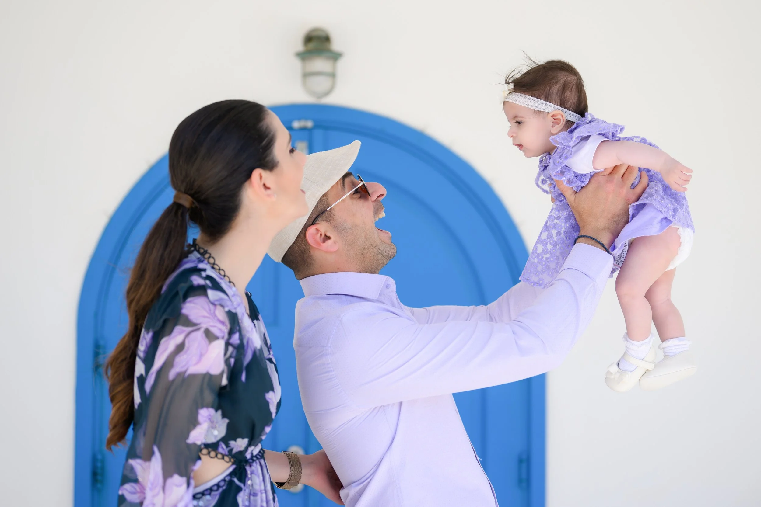 A man in a white shirt holding a young girl in a purple dress and headband up in the air with a woman beside them, all smiling.
