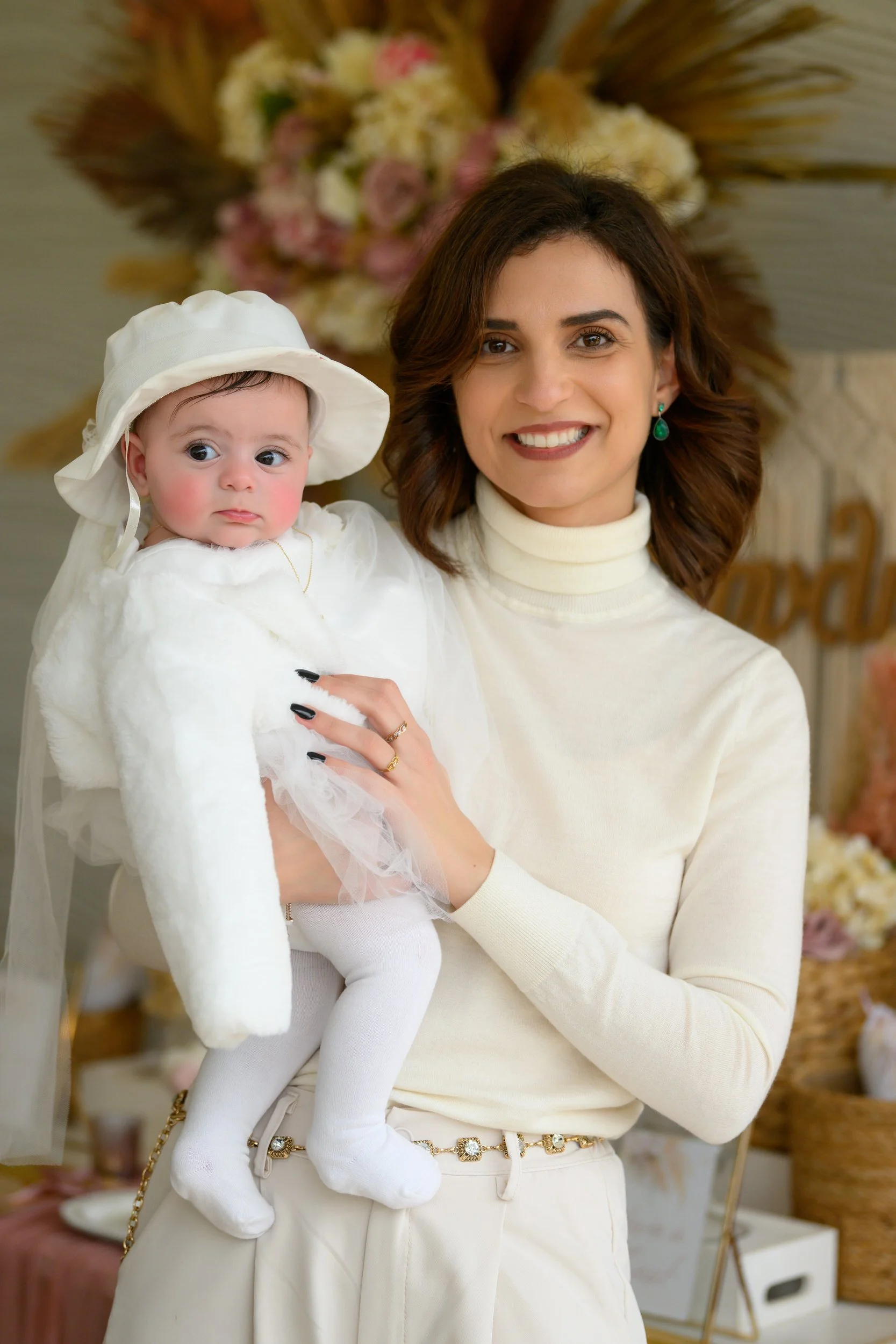 A woman holding a baby dressed in white, both smiling, with a floral arrangement in the background.