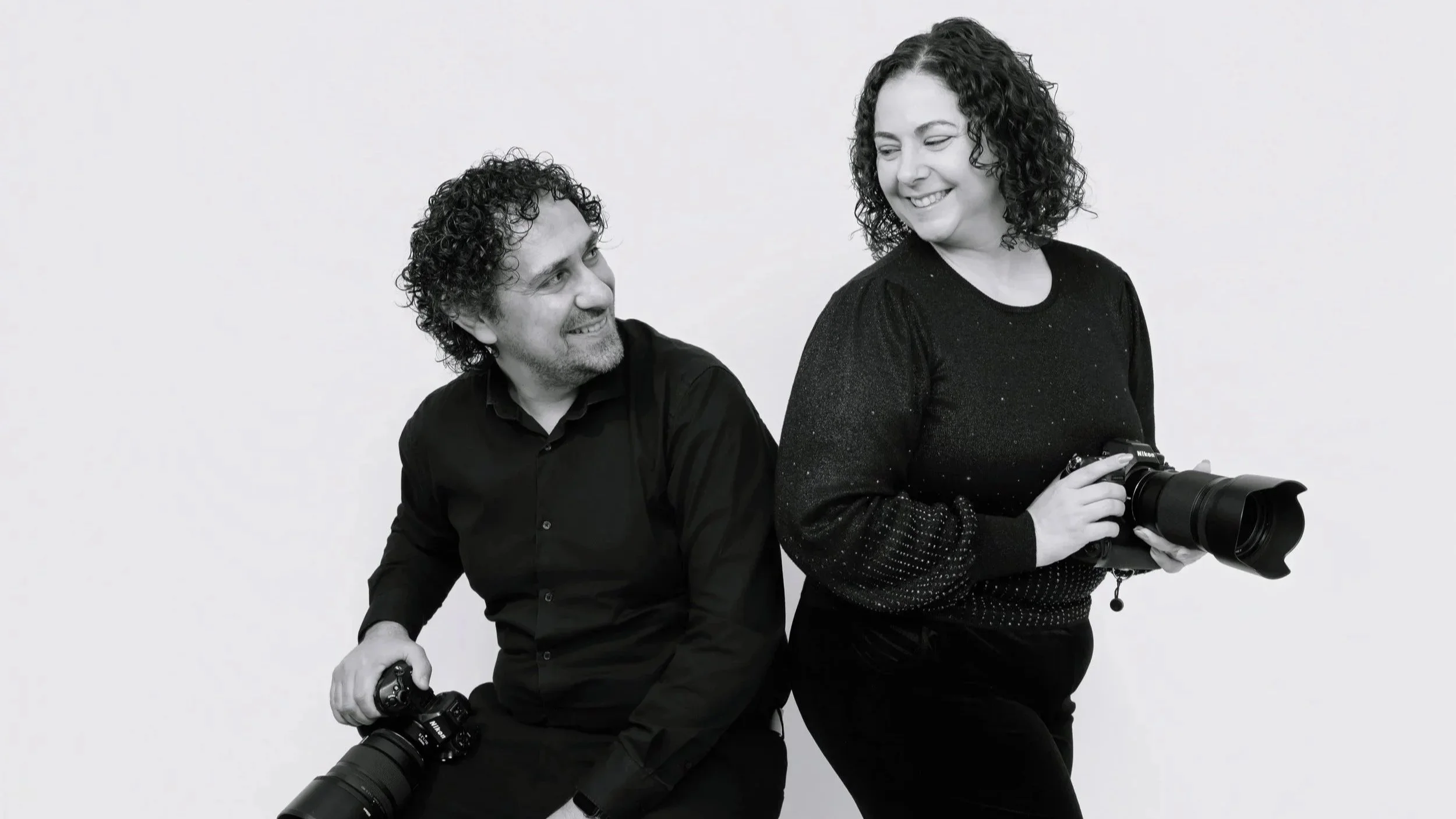 A man and a woman pose with cameras against a plain background, smiling at each other. The woman is holding a large camera in her hands, and the man is holding a camera with a telephoto lens, sitting on a stool. Both have curly hair and are dressed in dark clothing.