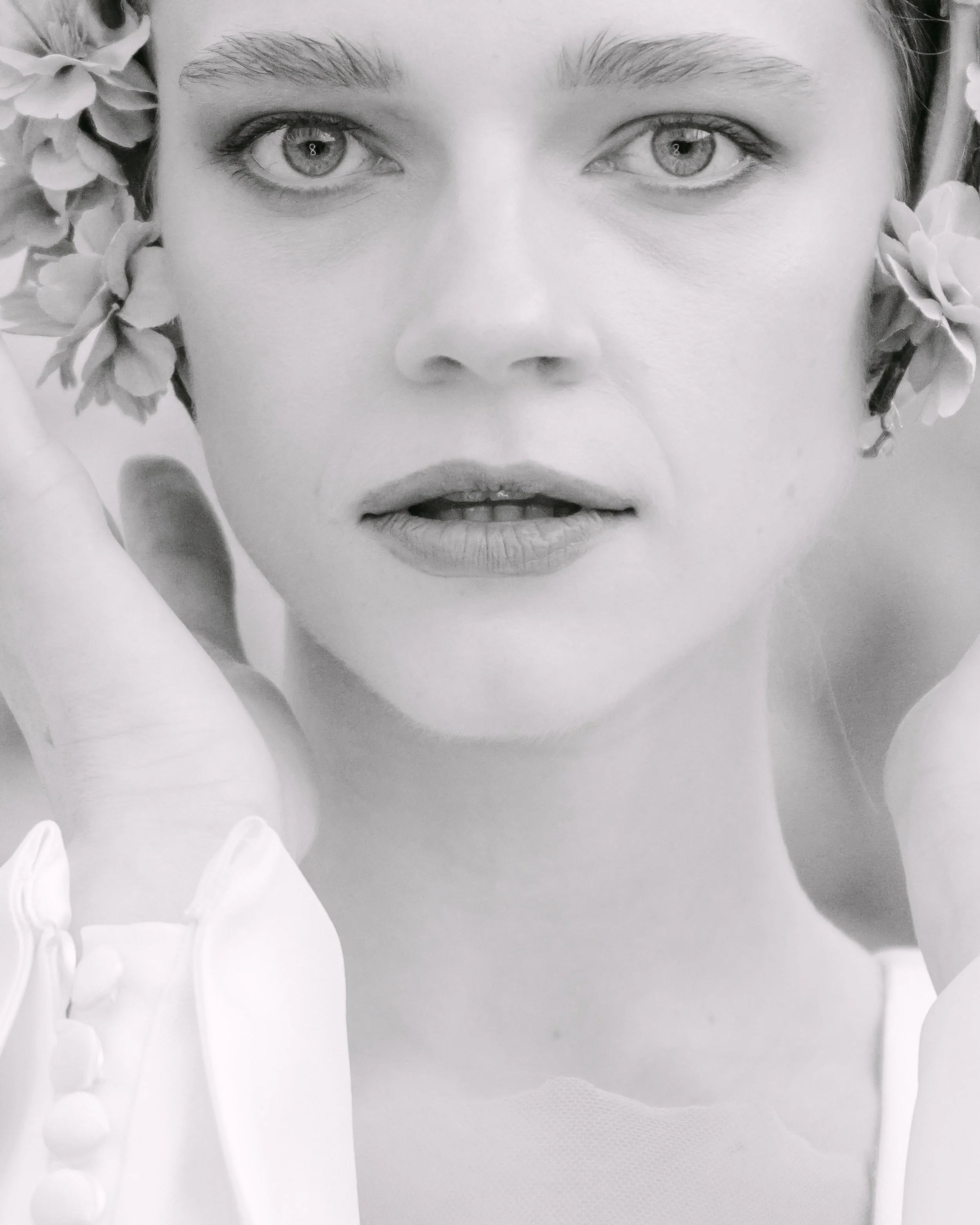 Black and white close-up portrait of a woman with flowers in her hair, holding her hands near her face with fingers gently touching her cheeks.