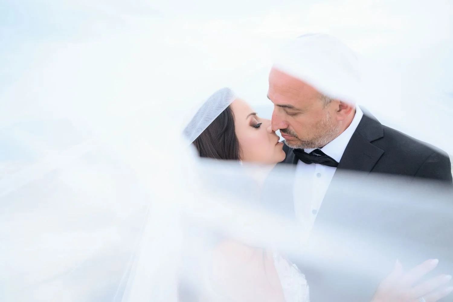 A bride and groom in wedding attire about to kiss, surrounded by a soft, blurred white veil or fabric.