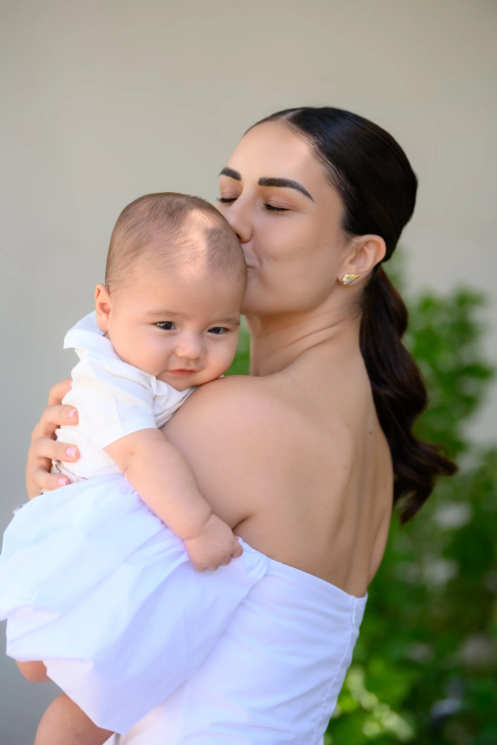 A woman with dark hair and gold earrings is holding a young child. She is gently kissing the child's forehead, who is gazing sideways. The background is out of focus with some greenery.