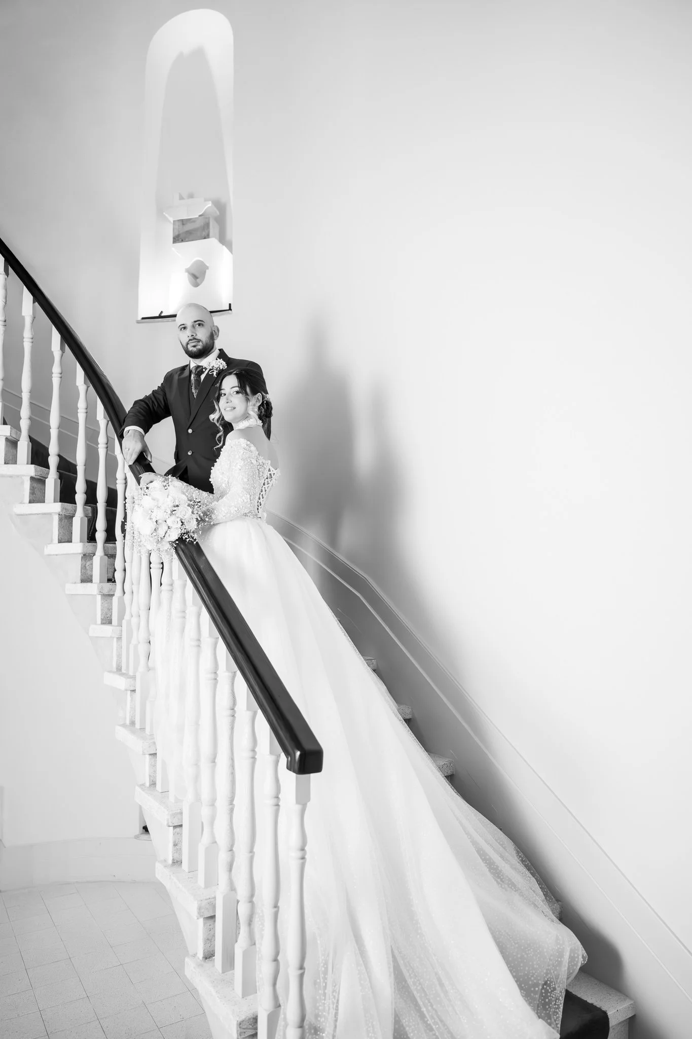 Black and white photo of a bride and groom standing on a staircase indoors, with the bride holding a bouquet of flowers and both looking at the camera.
