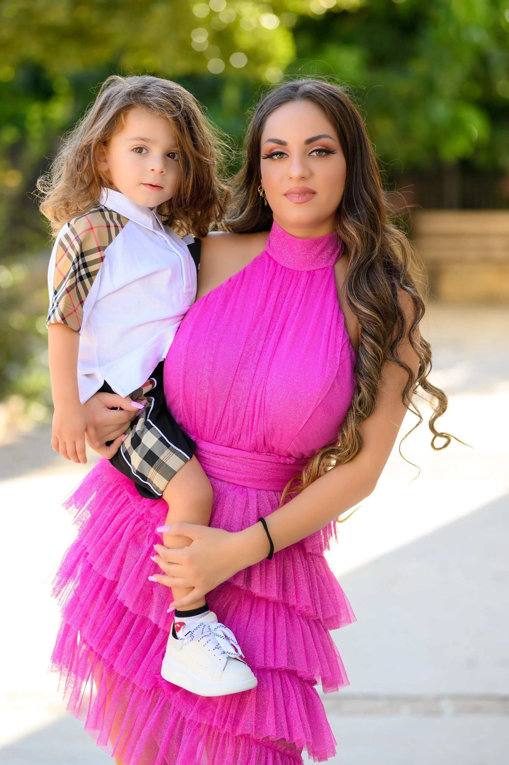 A woman with long wavy hair in a vibrant pink dress holding a young girl with light brown hair in a white and plaid outfit outdoors.
