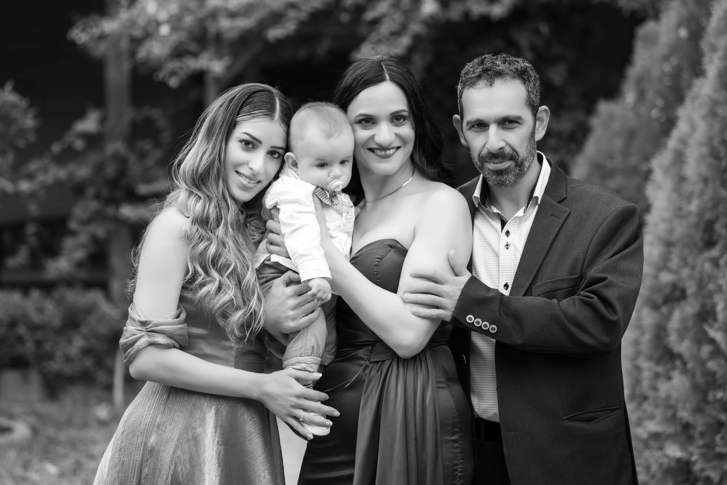 A black-and-white photo of a family of four outdoors, including a young woman with long wavy hair, a woman with dark hair holding a baby with a pacifier, an older man with a beard, all smiling and hugging each other.